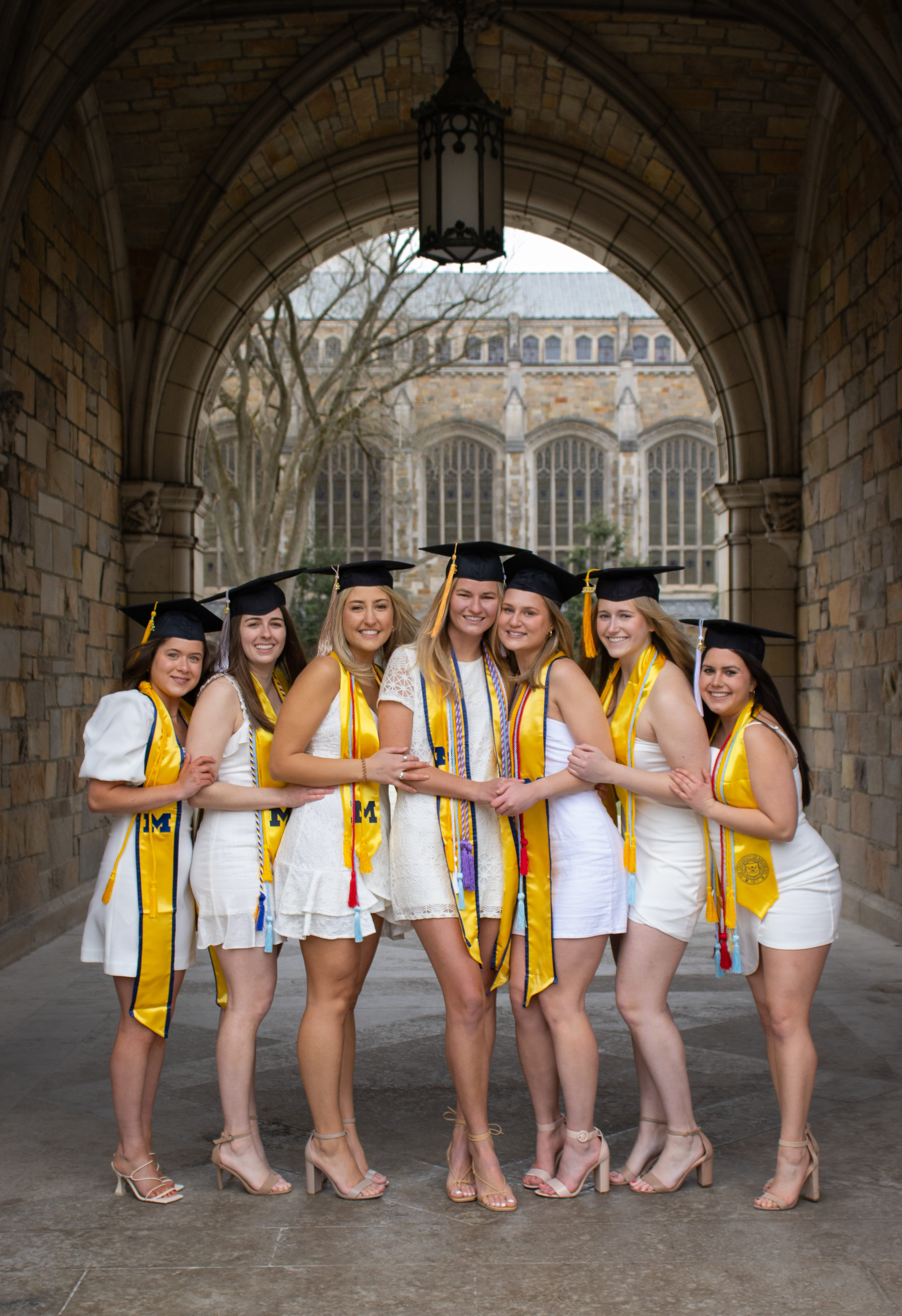 Seven female graduates in white dresses and caps with yellow stoles posing under stone archway.