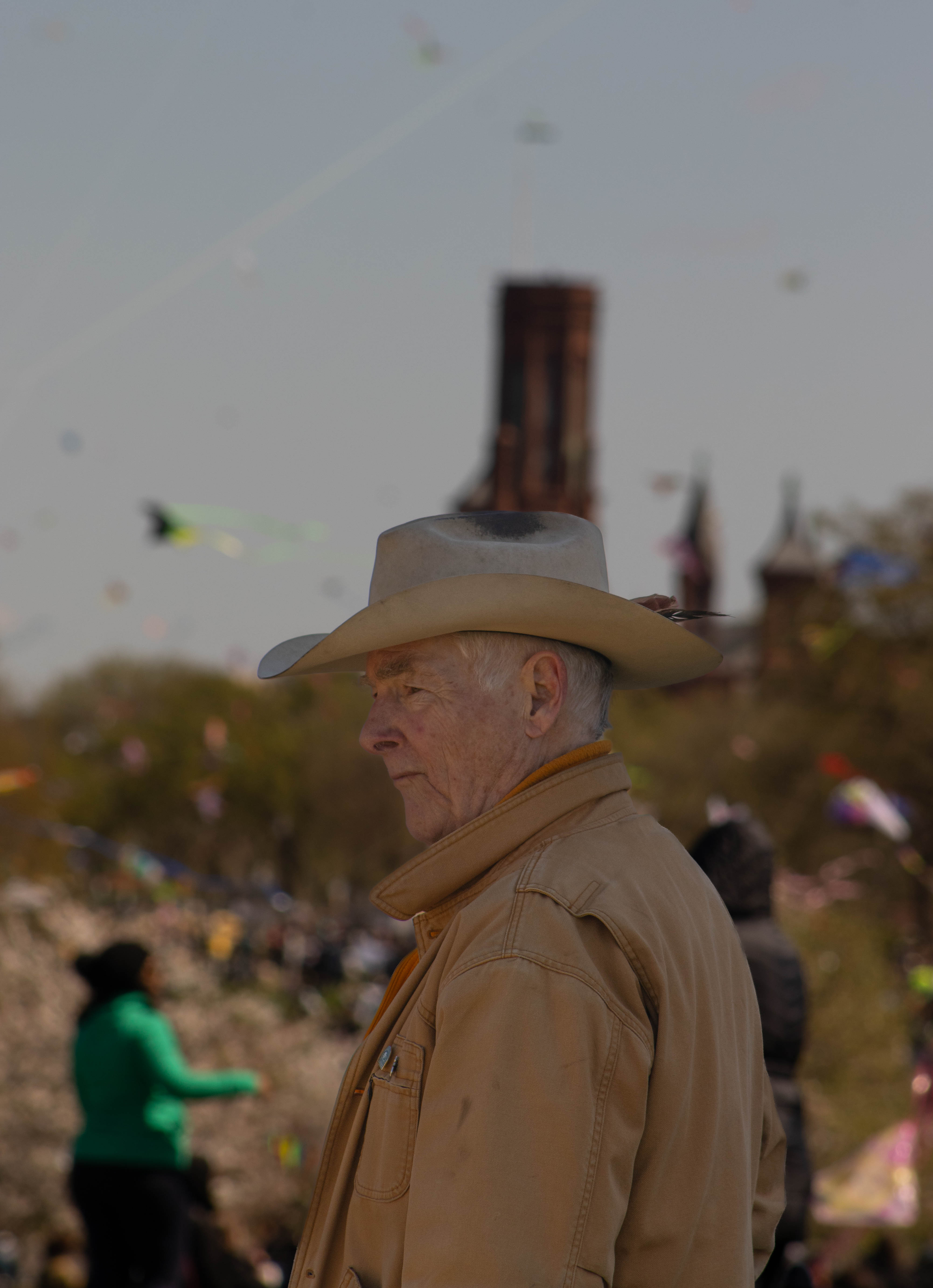 Elderly man wearing a beige cowboy hat and brown jacket, standing outdoors at a kite festival with kites flying in the background.
