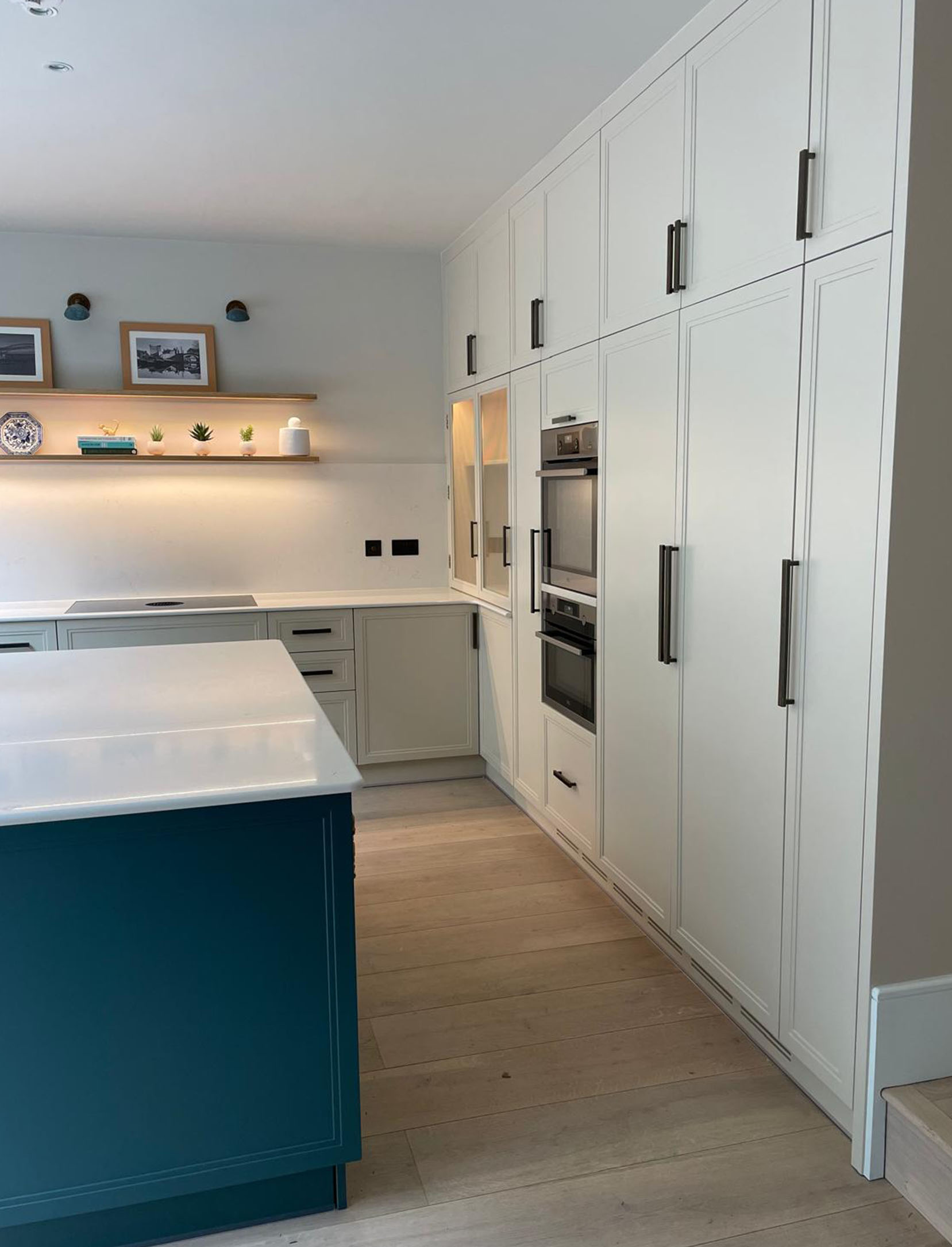 Integrated appliances sit seamlessly within the scheme, while illuminated glass-front display cabinets and open oak shelving with warm under-lighting introduce depth and softness. At the centre, a large L-shaped island with a crisp white quartz worktop anchors the space, combining generous preparation space with a calm, architectural presence. A dedicated drinks cabinet is concealed behind bifold doors with reeded glass, offering both function and a refined point of interest when opened. The island, finished in a contrasting tone, highlights the craftsmanship of its stepped panelling, adding quiet definition to the overall composition.