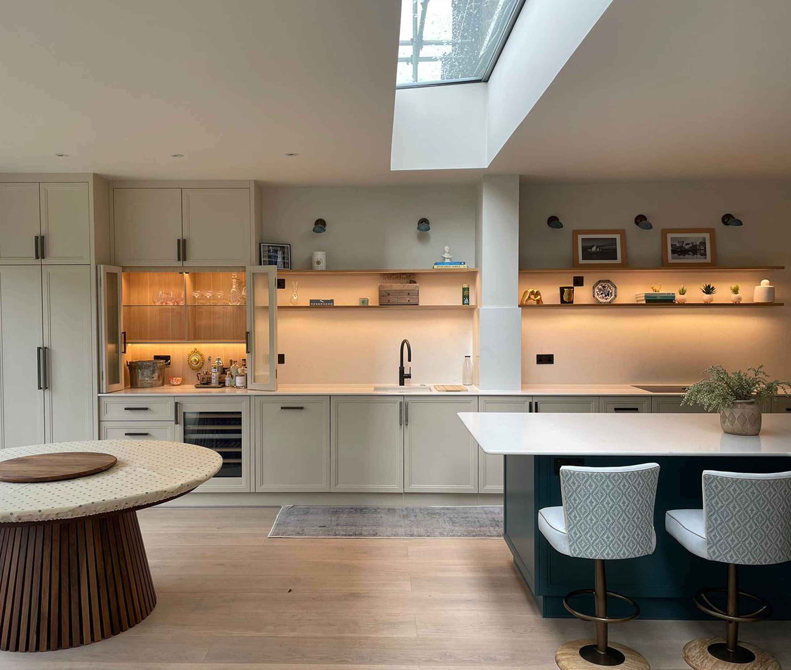 Integrated appliances sit seamlessly within the scheme, while illuminated glass-front display cabinets and open oak shelving with warm under-lighting introduce depth and softness. At the centre, a large L-shaped island with a crisp white quartz worktop anchors the space, combining generous preparation space with a calm, architectural presence. A dedicated drinks cabinet is concealed behind bifold doors with reeded glass, offering both function and a refined point of interest when opened. The island, finished in a contrasting tone, highlights the craftsmanship of its stepped panelling, adding quiet definition to the overall composition.