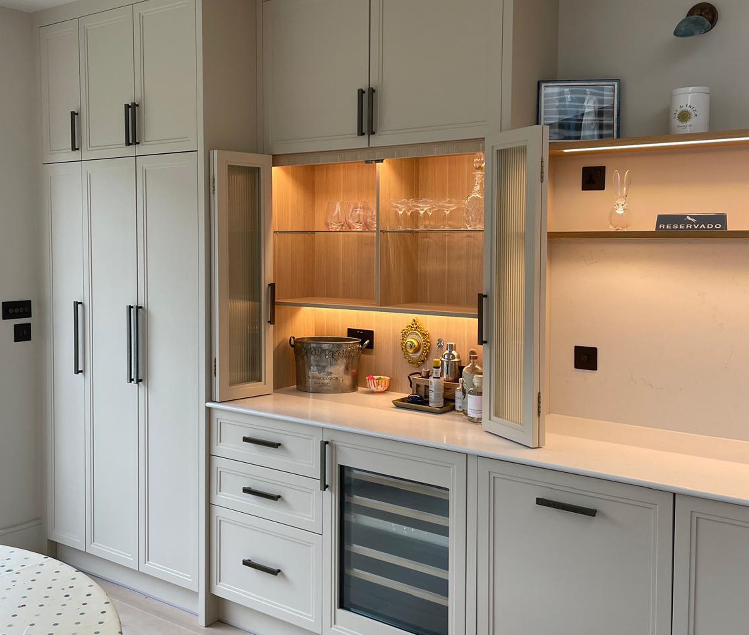 Integrated appliances sit seamlessly within the scheme, while illuminated glass-front display cabinets and open oak shelving with warm under-lighting introduce depth and softness. At the centre, a large L-shaped island with a crisp white quartz worktop anchors the space, combining generous preparation space with a calm, architectural presence. A dedicated drinks cabinet is concealed behind bifold doors with reeded glass, offering both function and a refined point of interest when opened. The island, finished in a contrasting tone, highlights the craftsmanship of its stepped panelling, adding quiet definition to the overall composition.