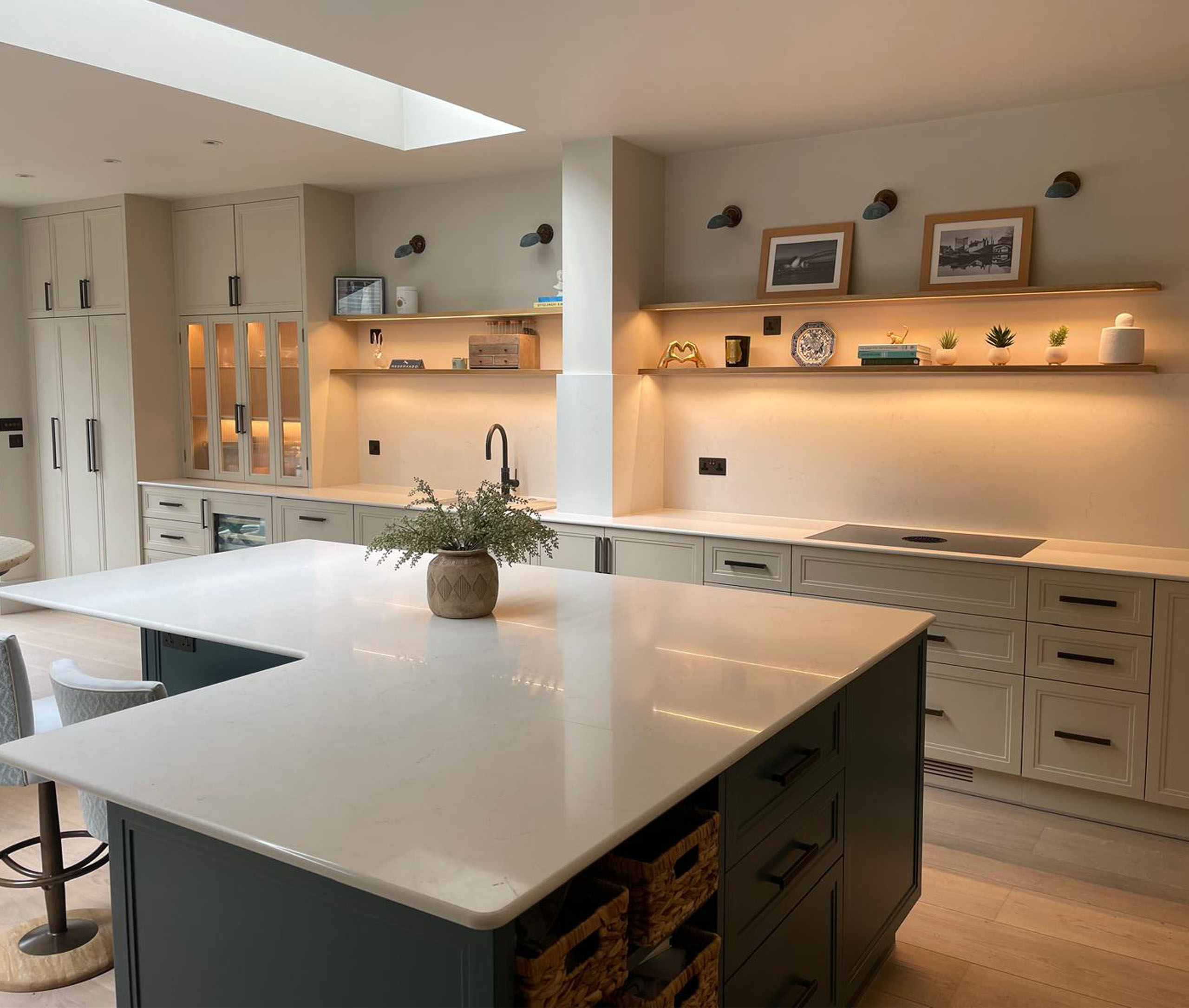 Integrated appliances sit seamlessly within the scheme, while illuminated glass-front display cabinets and open oak shelving with warm under-lighting introduce depth and softness. At the centre, a large L-shaped island with a crisp white quartz worktop anchors the space, combining generous preparation space with a calm, architectural presence. A dedicated drinks cabinet is concealed behind bifold doors with reeded glass, offering both function and a refined point of interest when opened. The island, finished in a contrasting tone, highlights the craftsmanship of its stepped panelling, adding quiet definition to the overall composition.