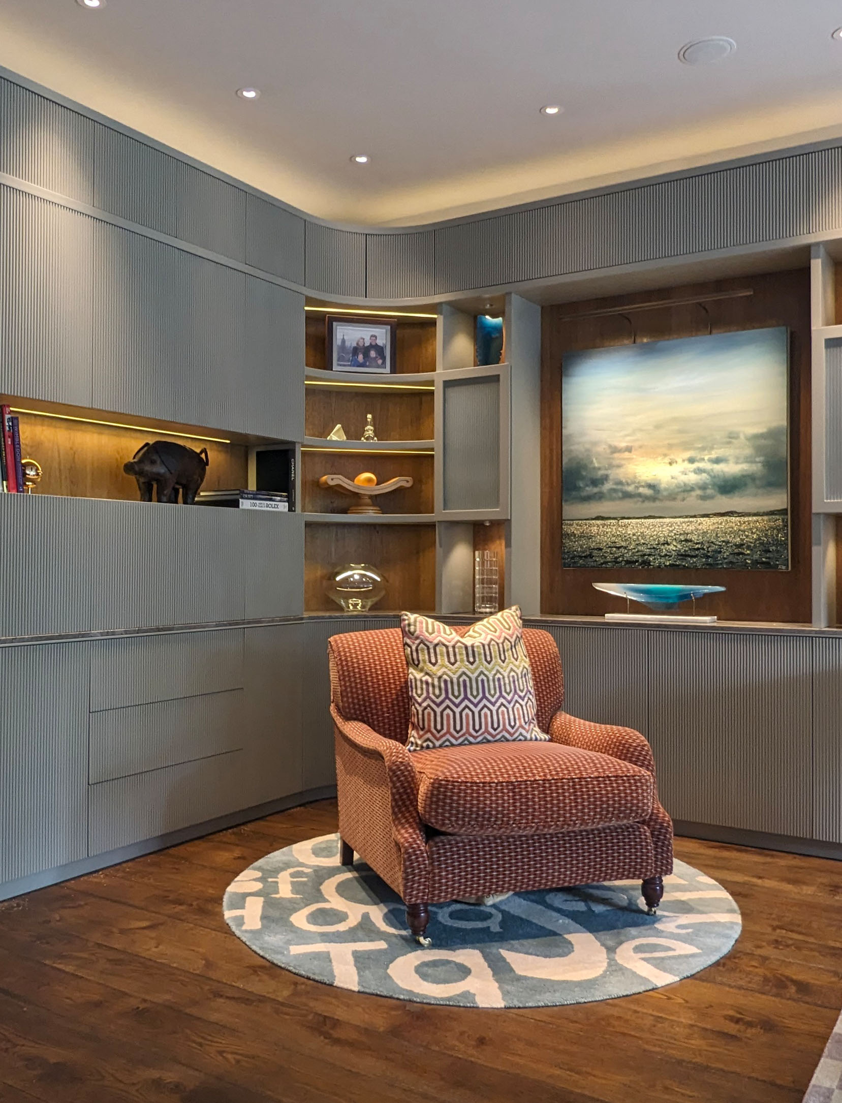 Cozy living room corner with patterned armchair on a round rug featuring abstract letters, surrounded by built-in gray shelving and a large seascape painting.