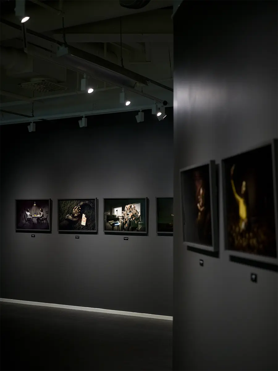 Mörk utställningshall med flera konstverk hängande i rad på en mörk vägg, upplyst av strålkastare.