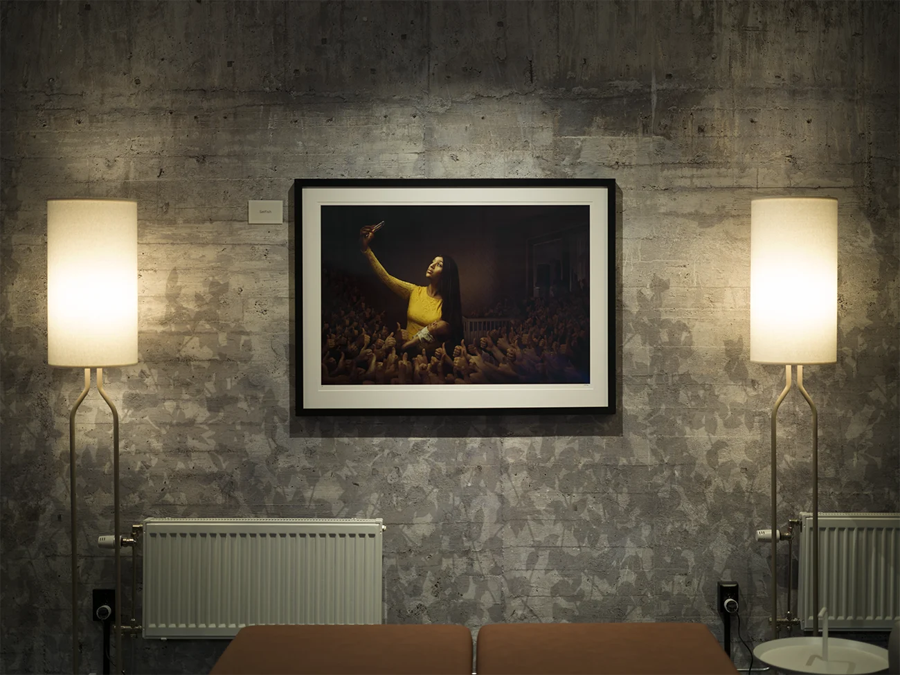 Konstverket Selfish hänger på en vägg mellan två lampor med en industrivägg.