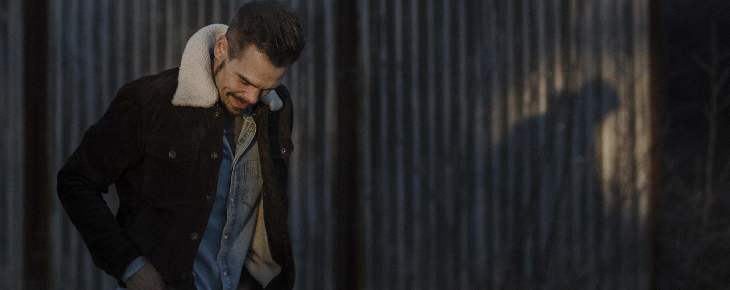 Artist standing in front of a steel wall, looking down, in background you can see his silhouette and shadow on the wall. 