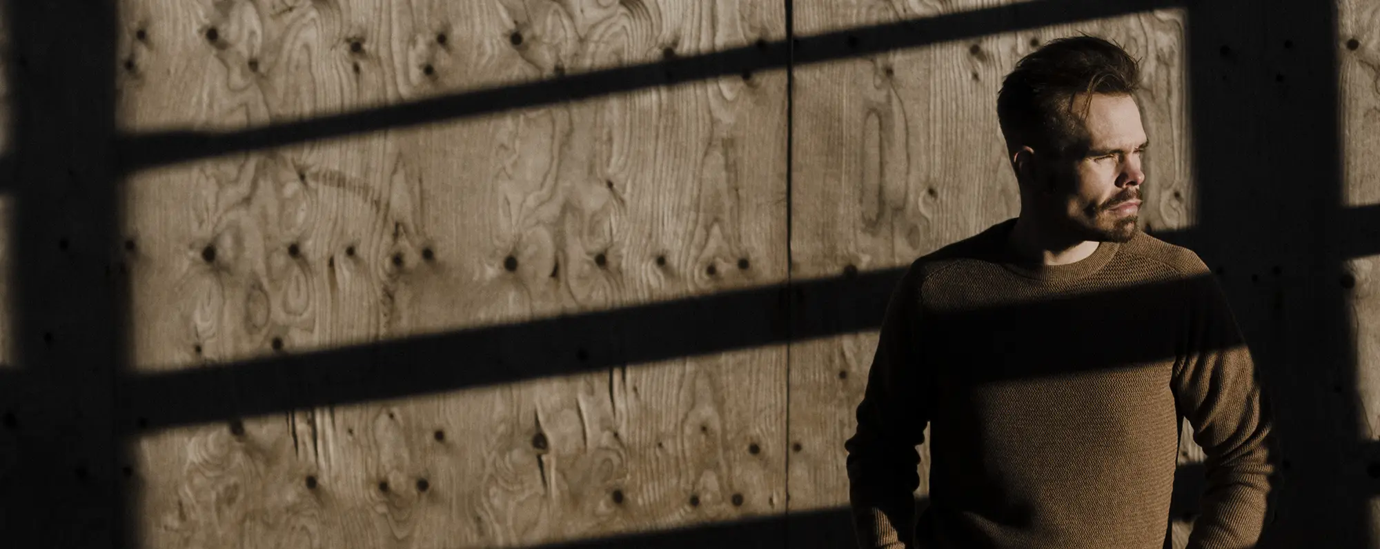 Man standing in front of a wooden wall, looking to his right into the sunlight, squinting with his eyes.