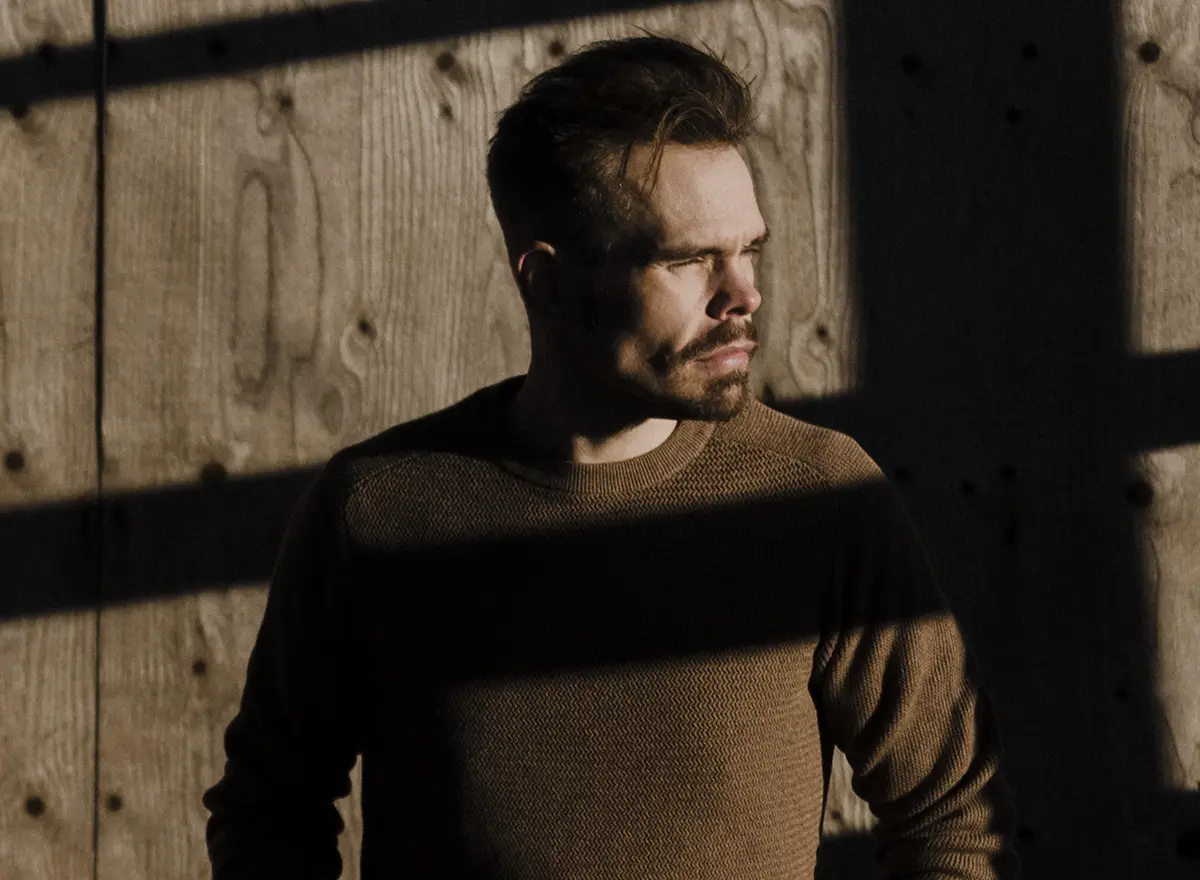 Man standing in front of a wooden wall, looking to his right into the sunlight, squinting with his eyes.