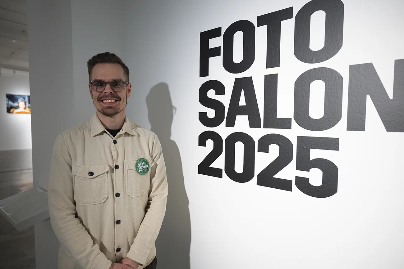 Andreas Varro poserar framför en väggskylt som säger FOTO SALOON 2025