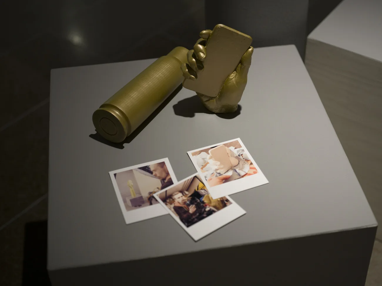 Rekvisita som ligger på ett bord i en utställningshall. En hand som håller en telefon, ett kulskal och tre polaroidbilder.