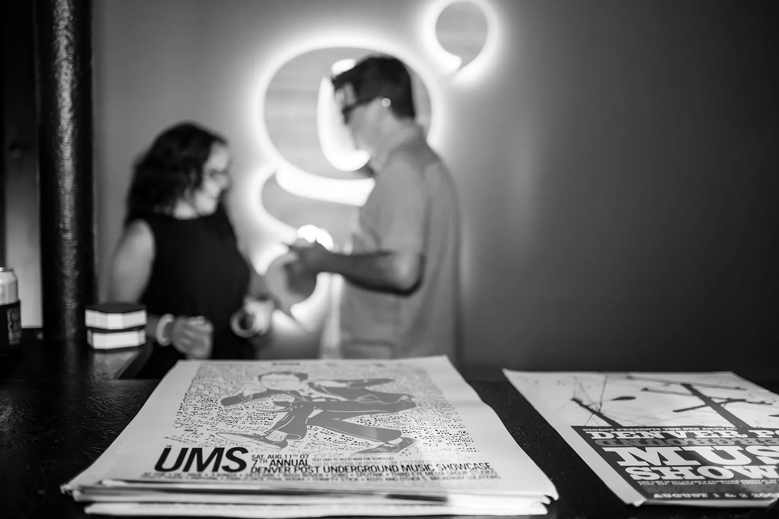 Black and white photo showing a stack of UMS music showcase flyers on a counter, with two blurred people talking in the background.