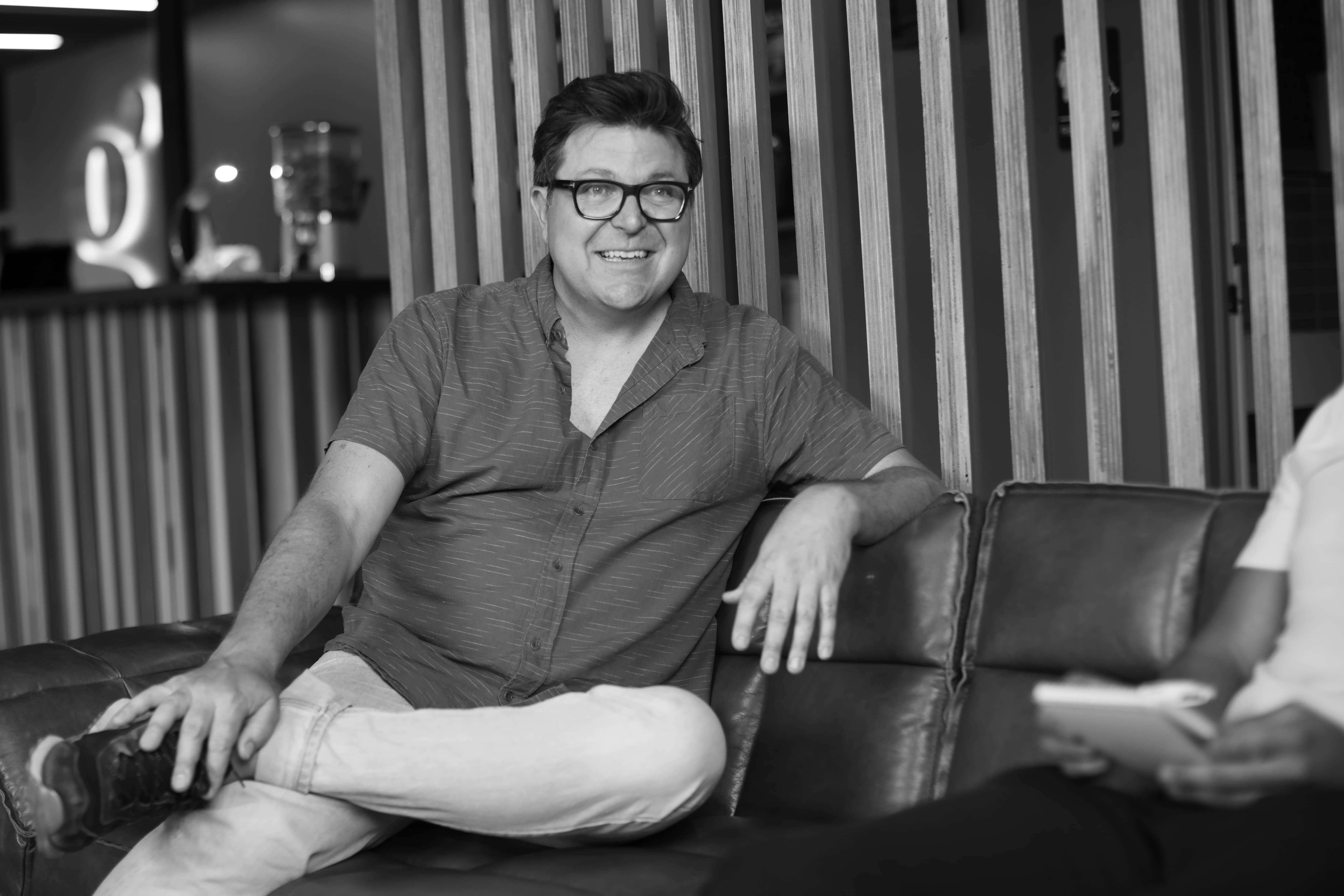 Smiling man wearing glasses and a short-sleeve shirt sitting cross-legged on a couch indoors.