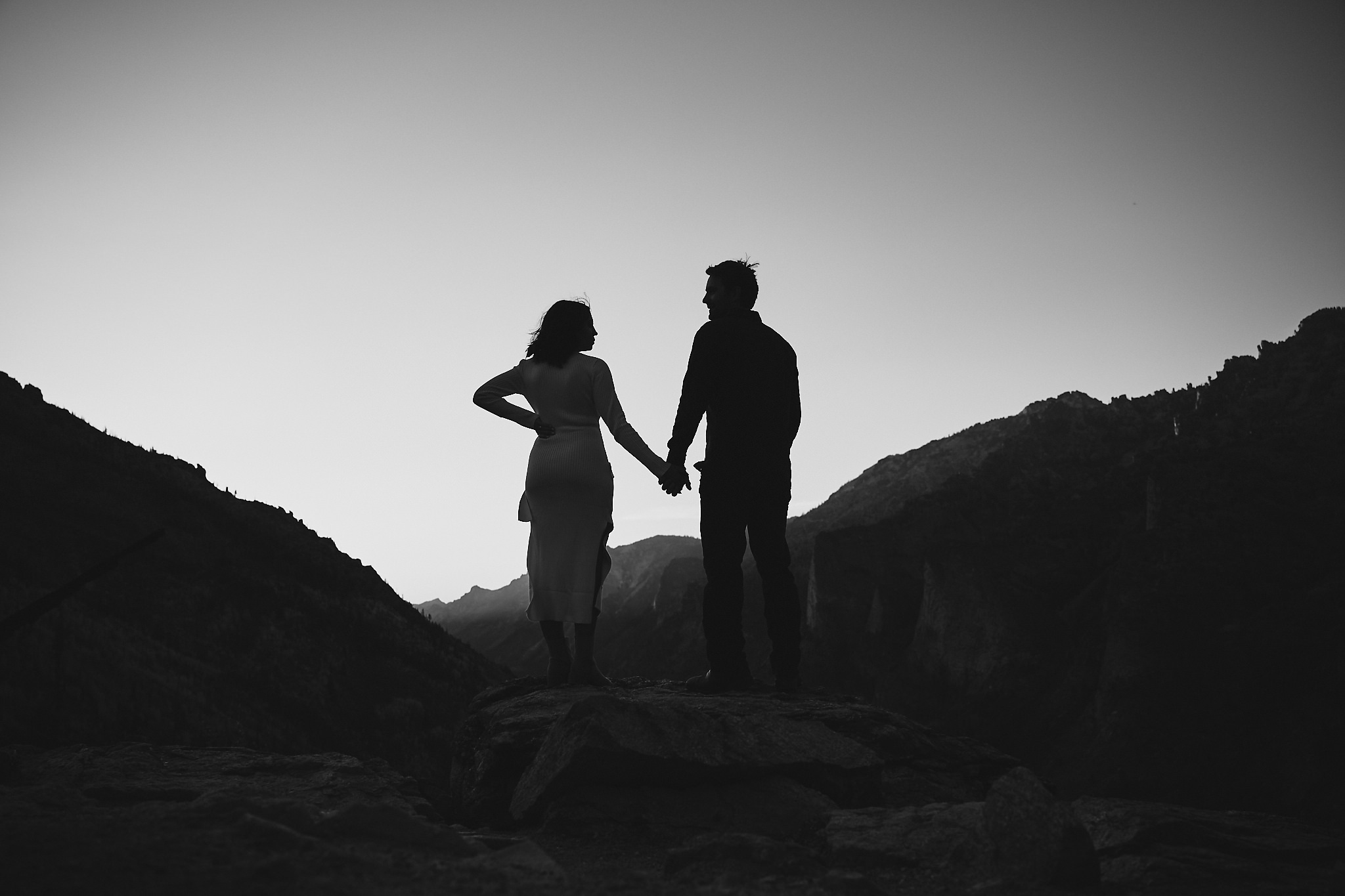 Silhouetted couple holding hands on a rock overlooking mountainous terrain at dusk.