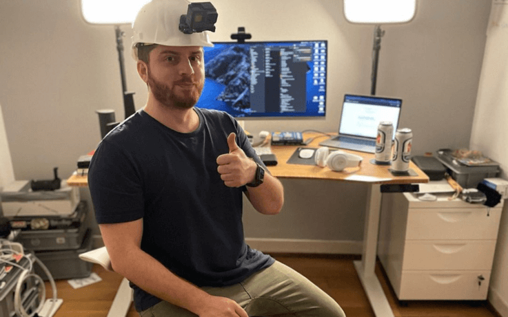 man sitting with safety hat and camera on forehead with desk and screens in the background