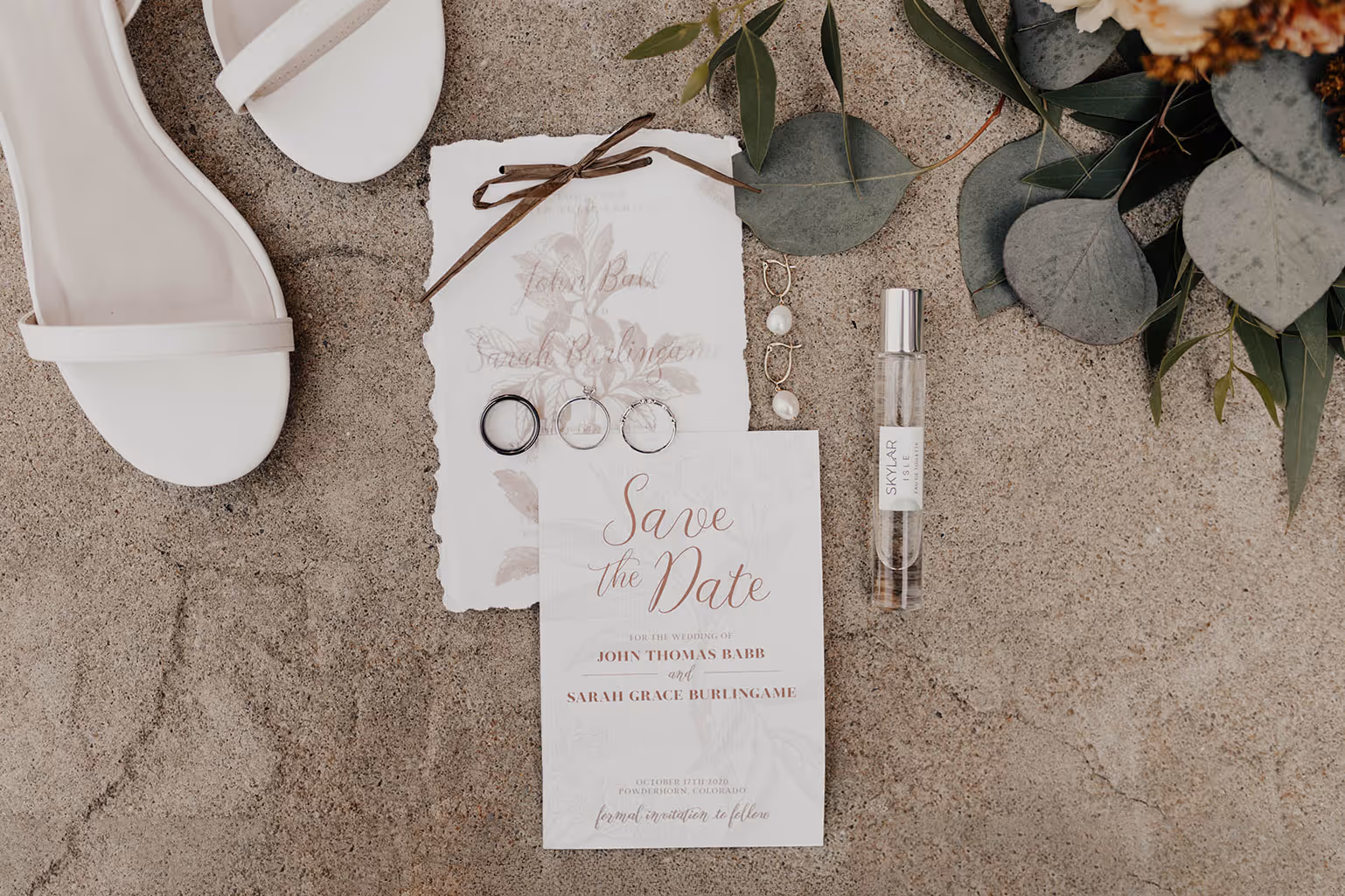 An invitation to a wedding laying on a stone surface, with white heels nearby, slim bottle of perfume, and delicate pearl earrings sit nearby. The invite is below a Save the Date card - both are floral in design with multiple layers and textures. A rafia ribbon ties a transluscent floral design to a thick laid deckle edge card stock.