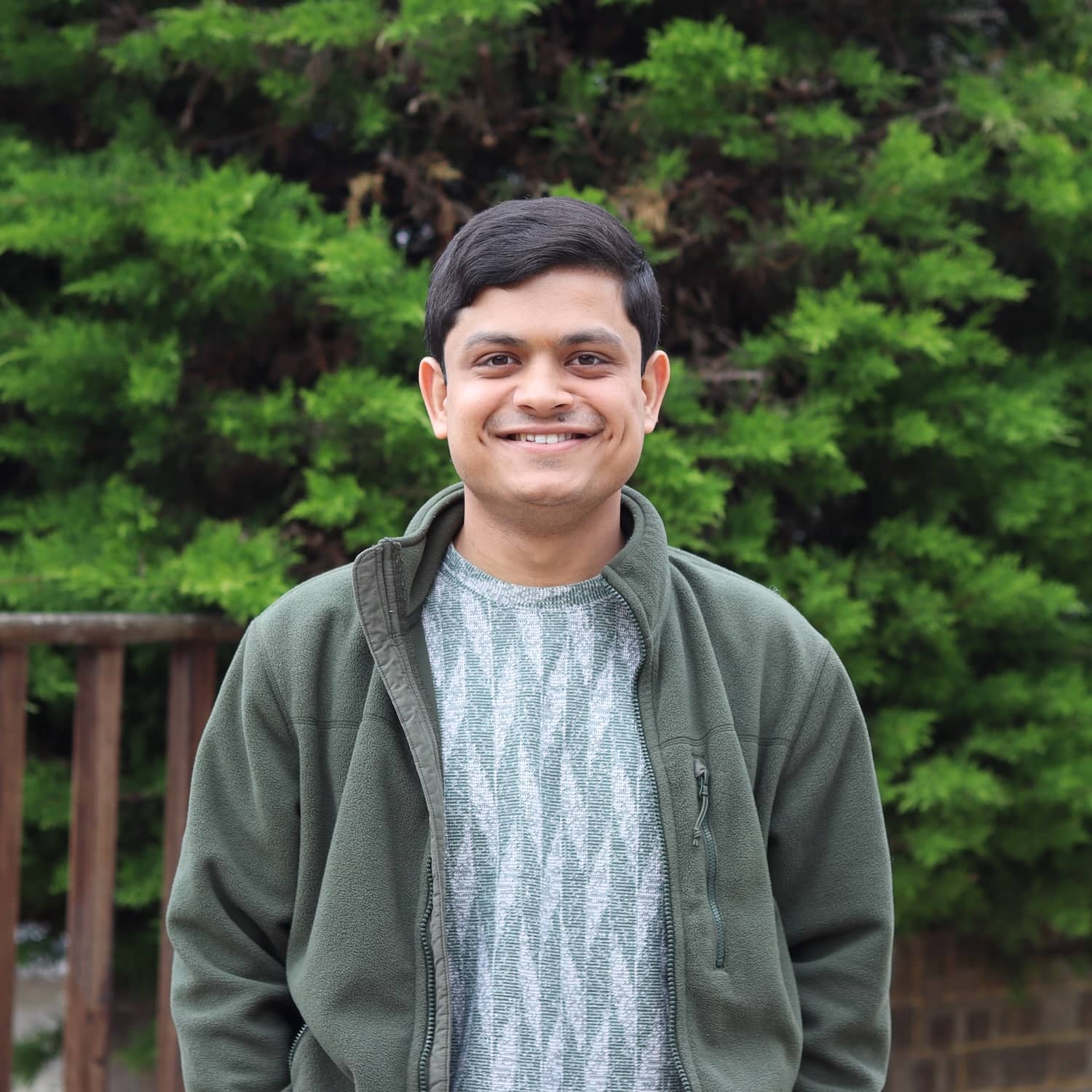 Image of a man with black hair from the waist up, smiling. With an evergreen tree in the background.