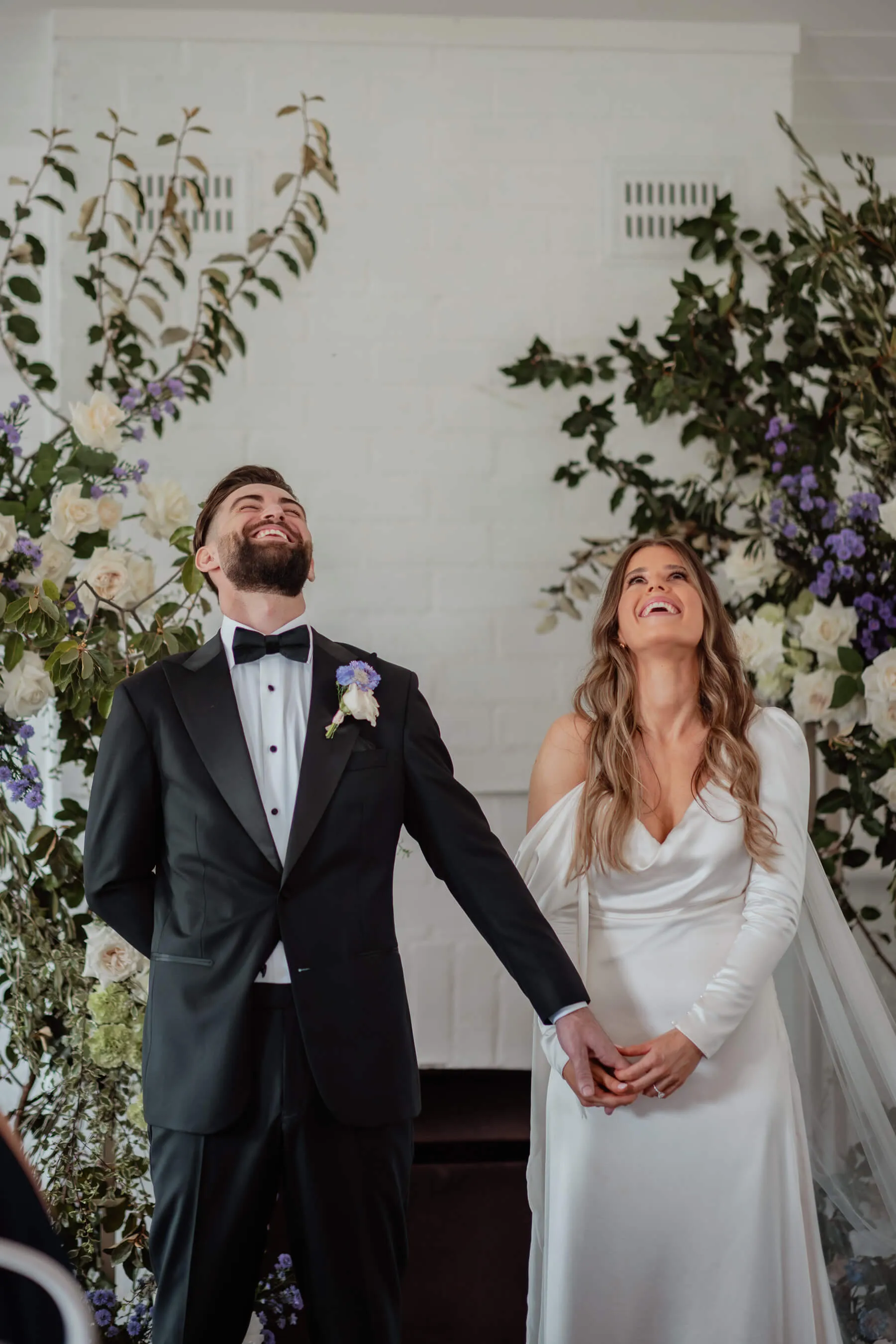Monica and Luke smiling beneath a floral arch during their outdoor wedding ceremony