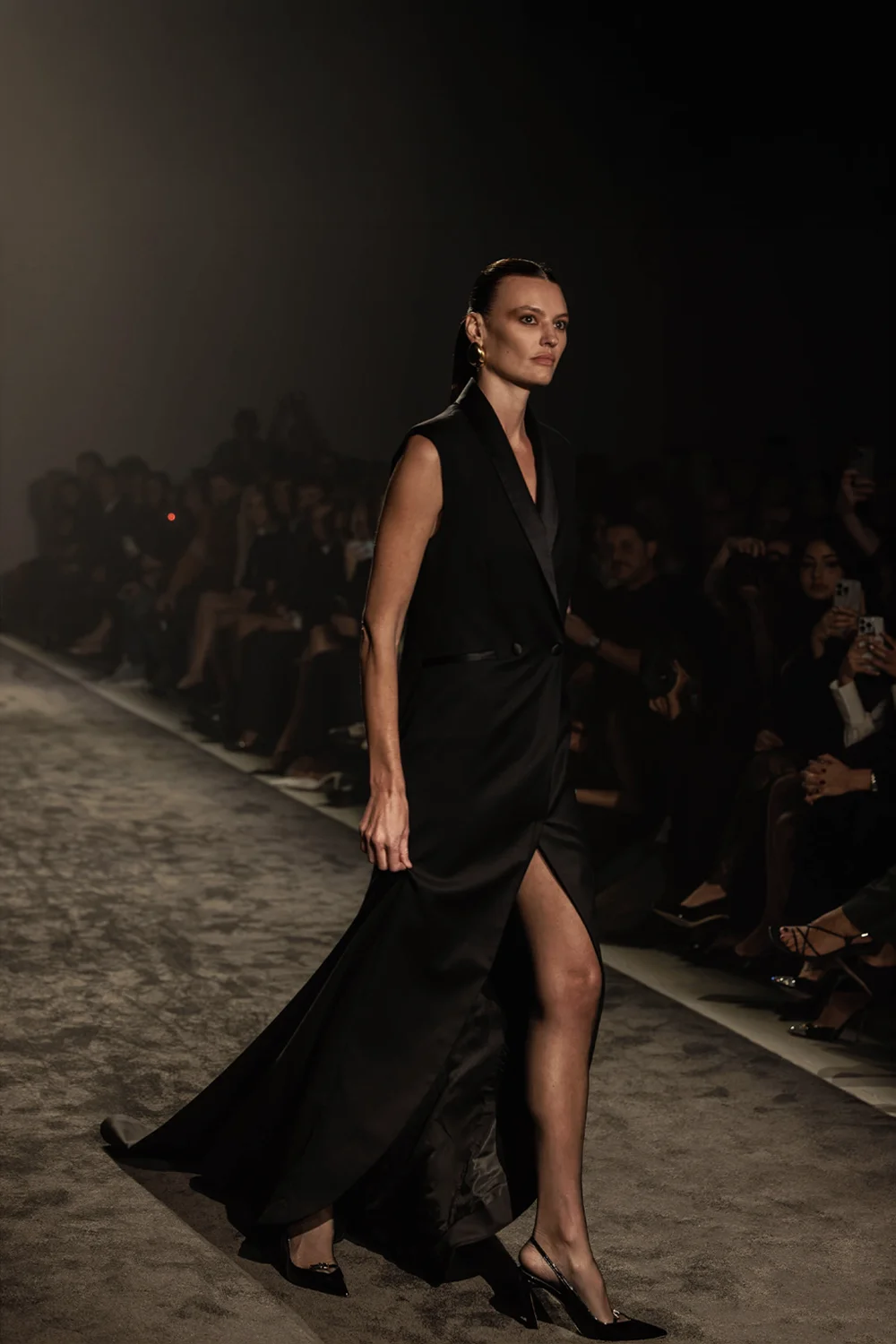 Side profile of a model in a floor-length black tuxedo gown with a dramatic slit and gold earrings on the Farage Australia Summer 26 runway at AFW 2025.