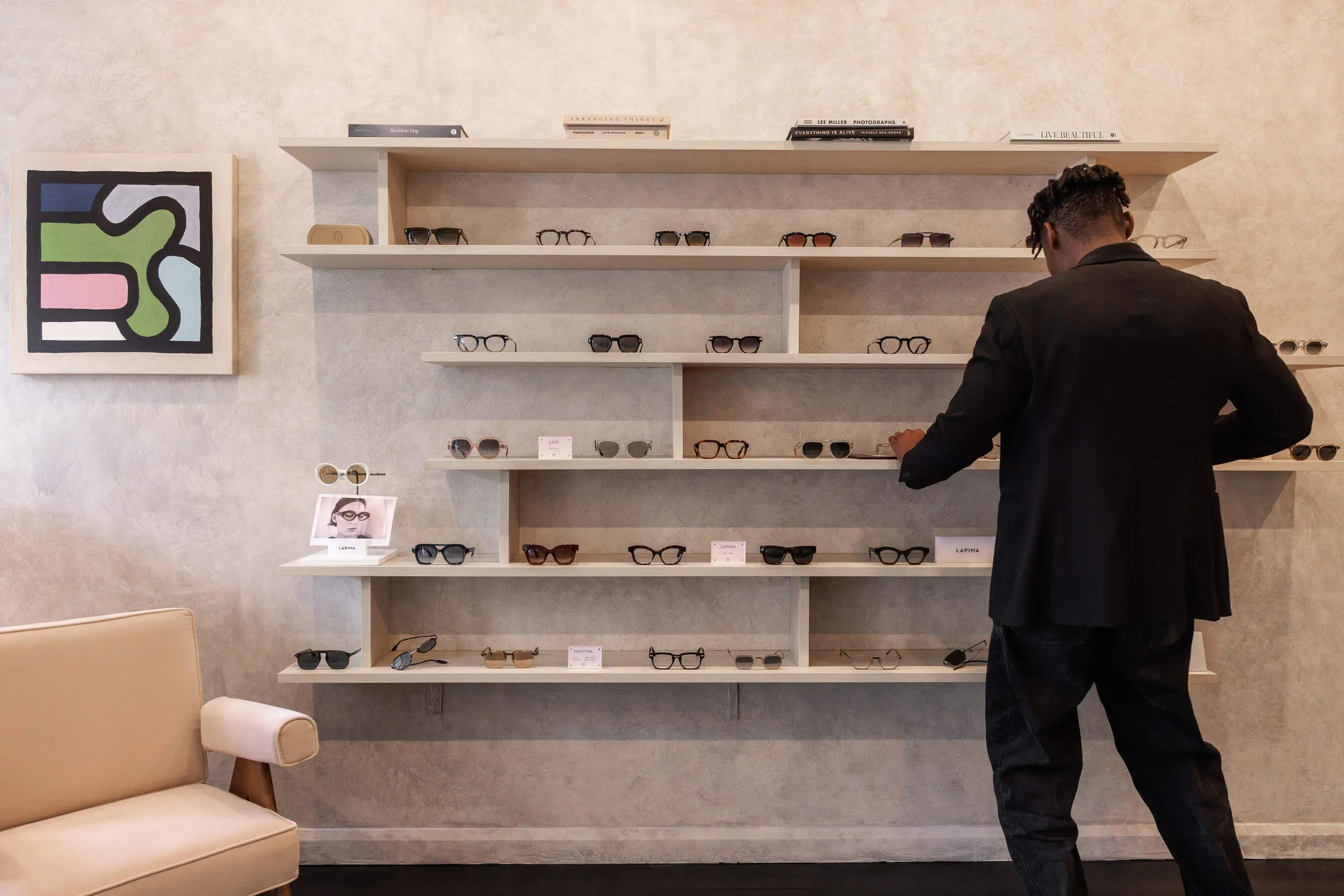 Candid in-store moment of a customer browsing the eyewear collection on wall-mounted shelves. The layout shows multiple designer brands including LAPIMA and DITA, captured against the textured feature wall | EDG Studios