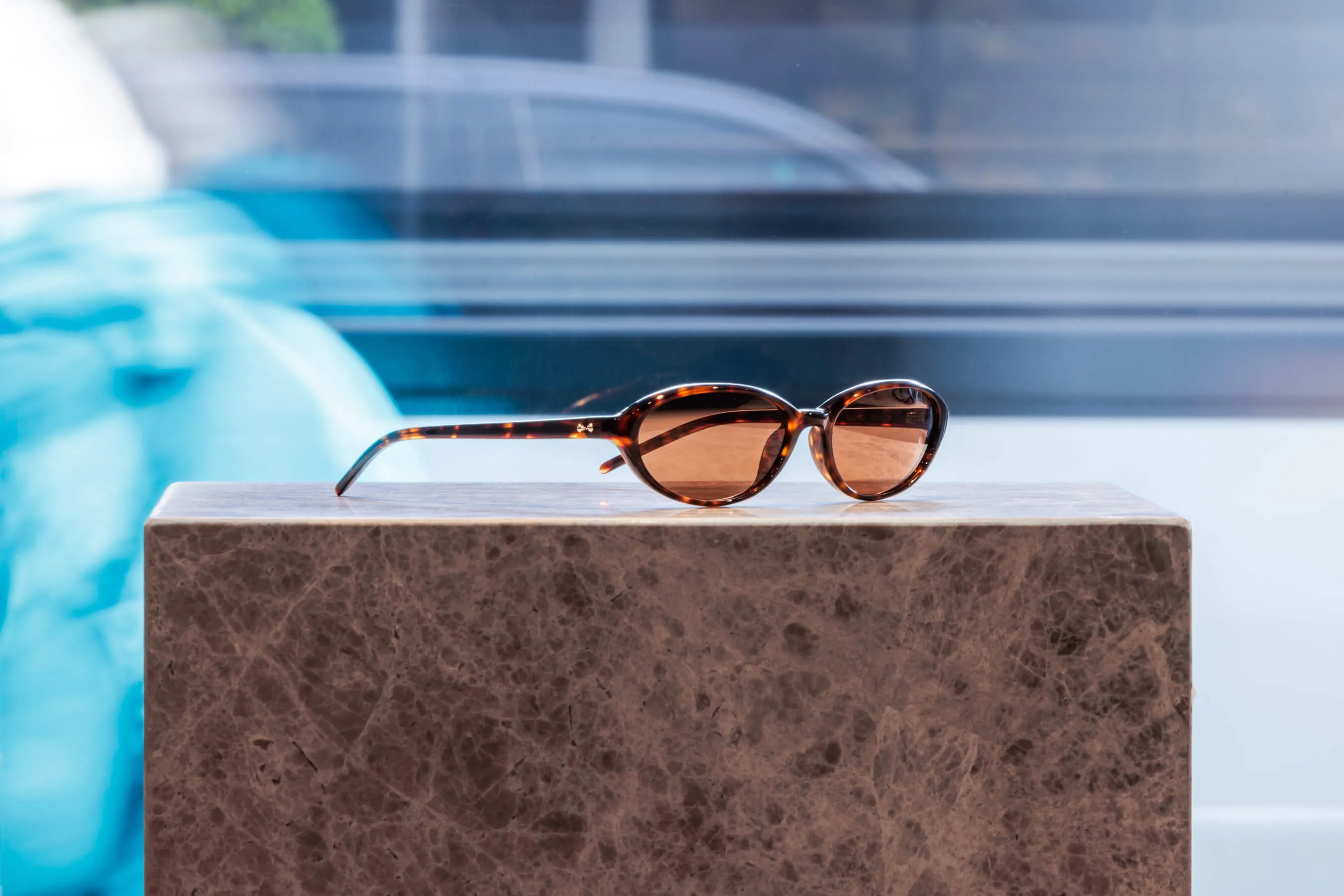 Product feature of oval tortoiseshell sunglasses with warm brown lenses, displayed on a polished marble plinth near the shopfront. A blurred street scene in the background adds subtle urban context | EDG Studios