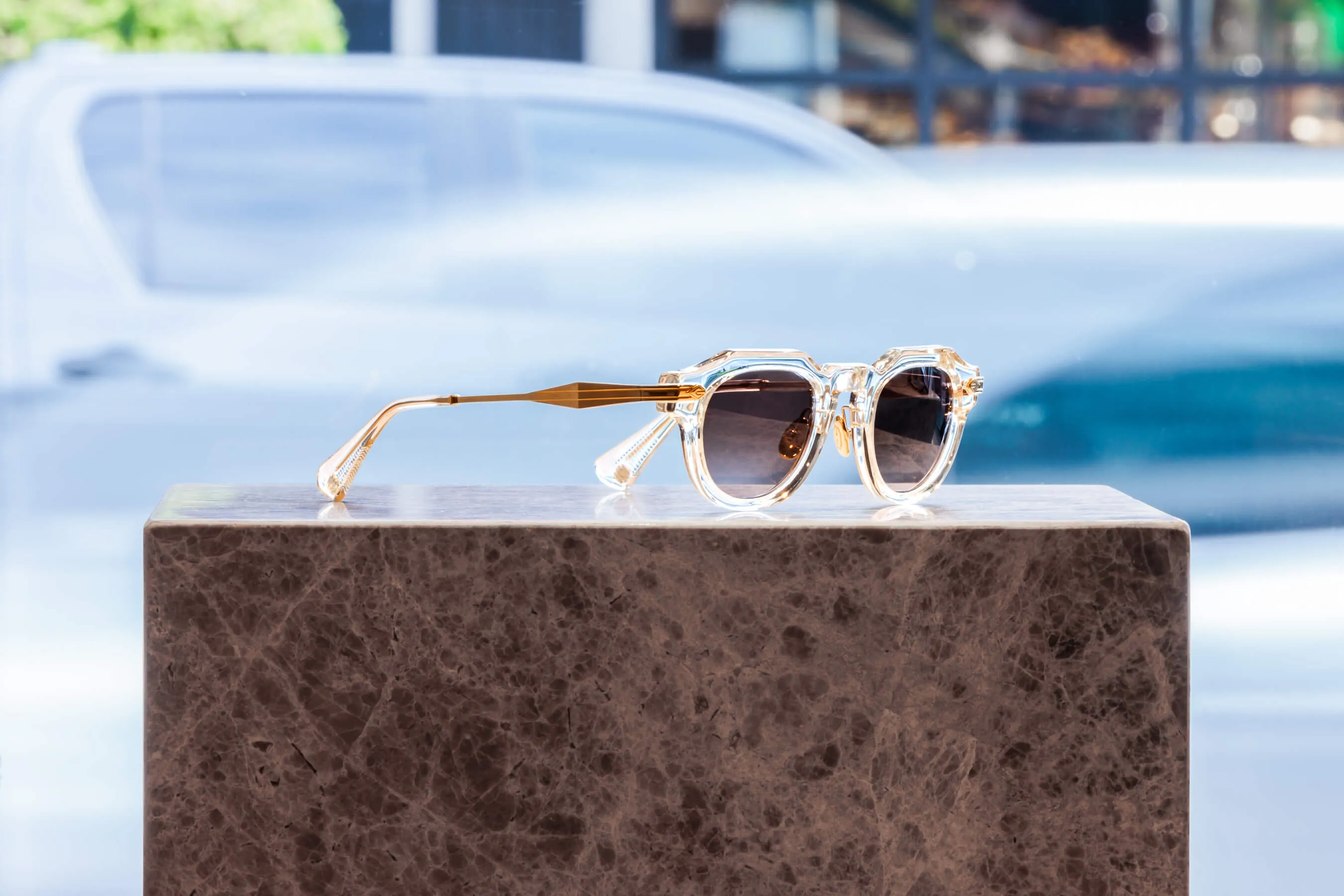 Crystal-clear acetate sunglasses with sculpted gold-tone arms rest on a marble plinth. Sunlight enhances the clarity of the frame and texture of the lenses, set against an out-of-focus vehicle | EDG Studios