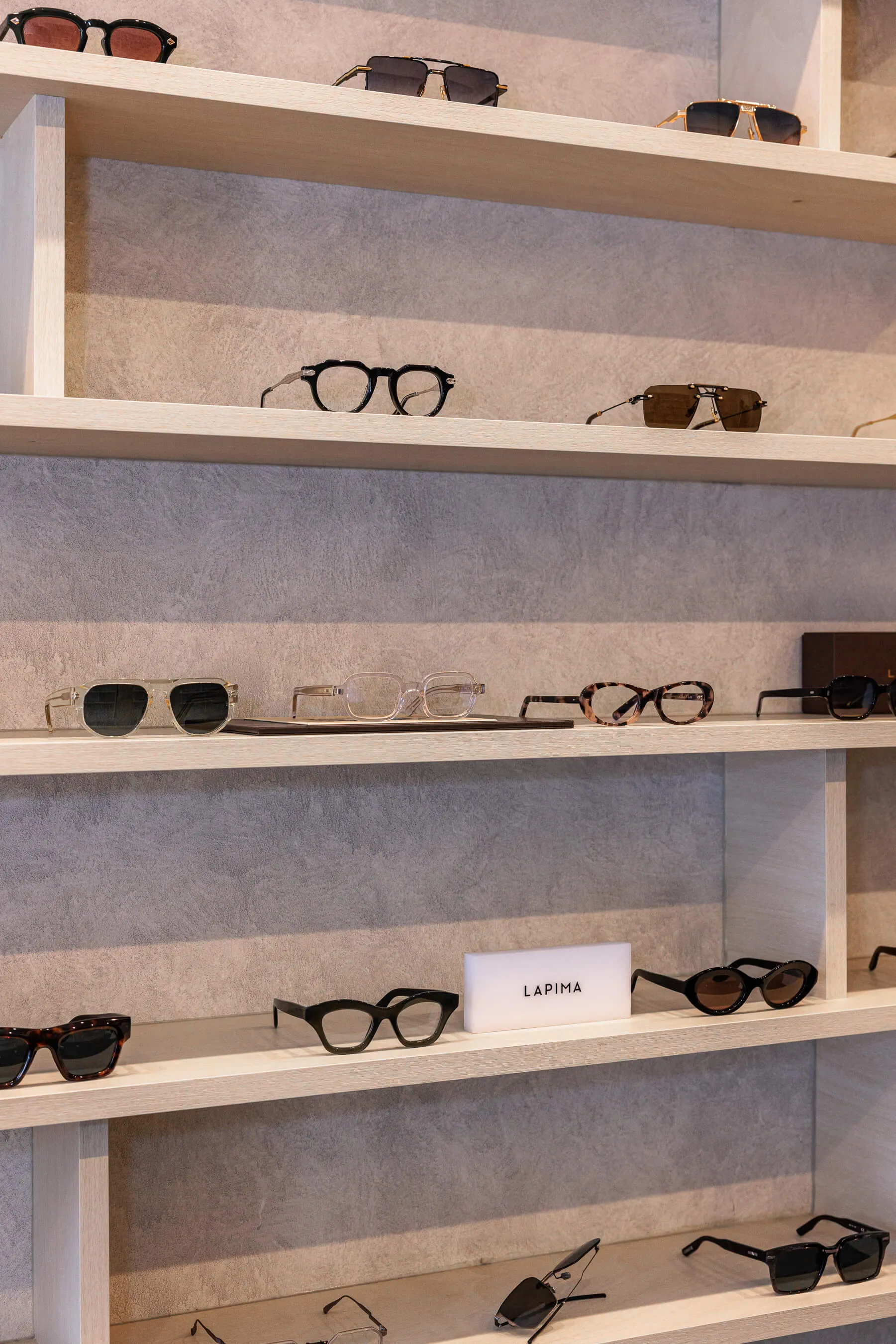 Close-up of multiple LAPIMA eyewear styles displayed on pale wood shelving. A range of acetate frames in various colorways and shapes is neatly organized beneath minimalist branding | EDG Studios