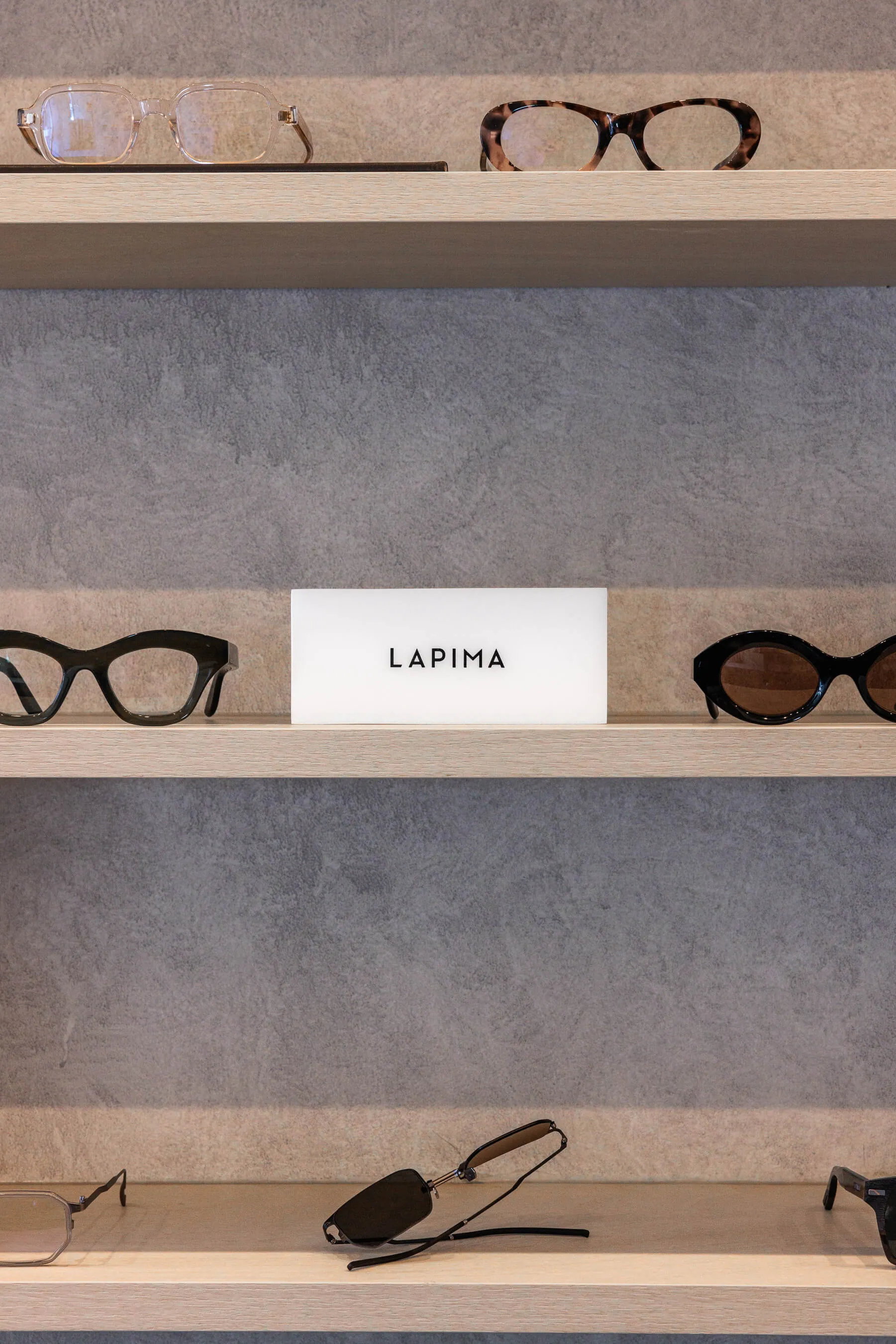 Focused image of the LAPIMA brand plaque flanked by distinctive sunglasses on either side. Matte black and tortoiseshell frames contrast against the cool-toned, concrete-textured wall | EDG Studios