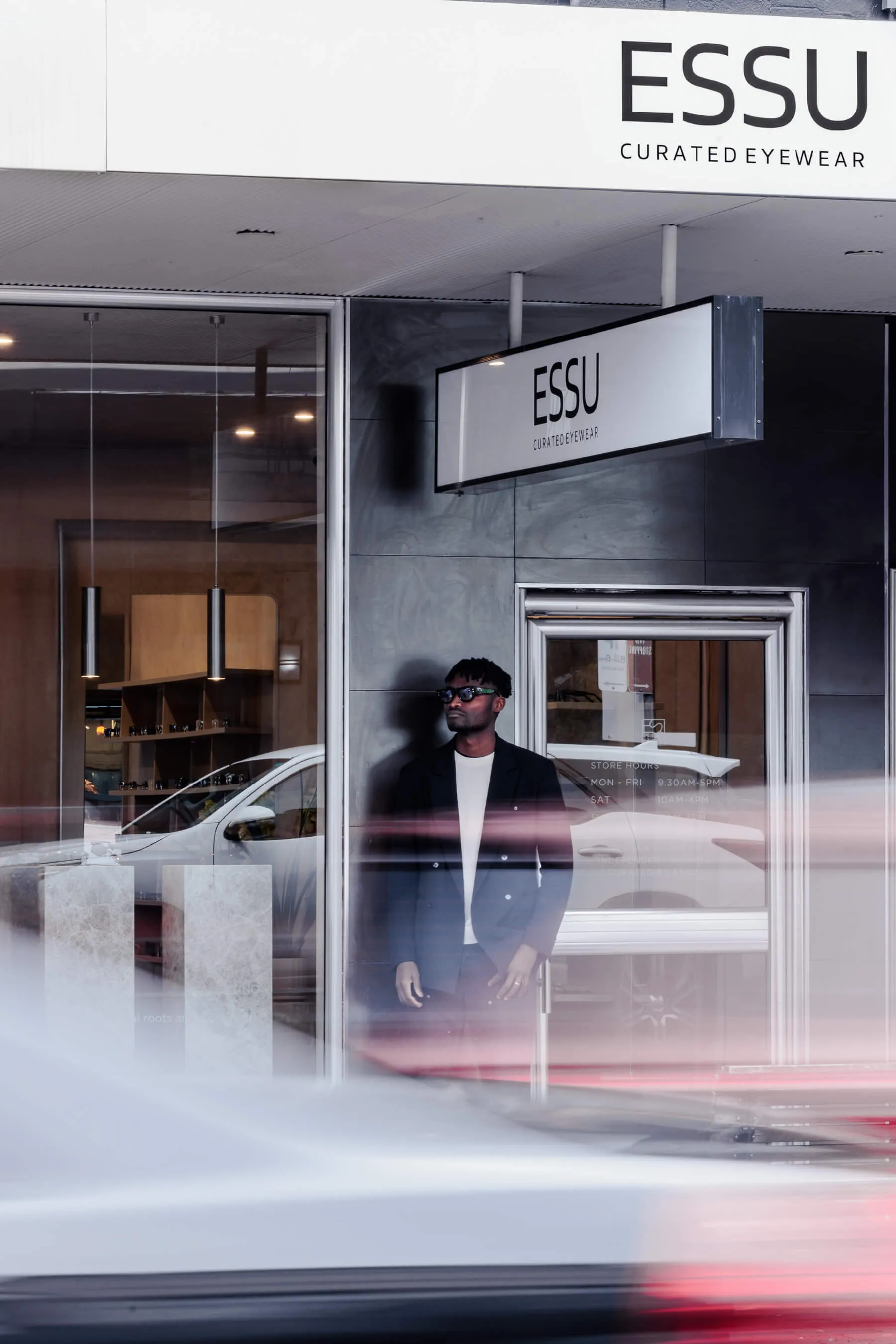 Street view of the Essu storefront. A sharply dressed model wearing sunglasses stands under the ESSU signage, as streaks of passing traffic create a sense of movement and urban energy | EDG Studios