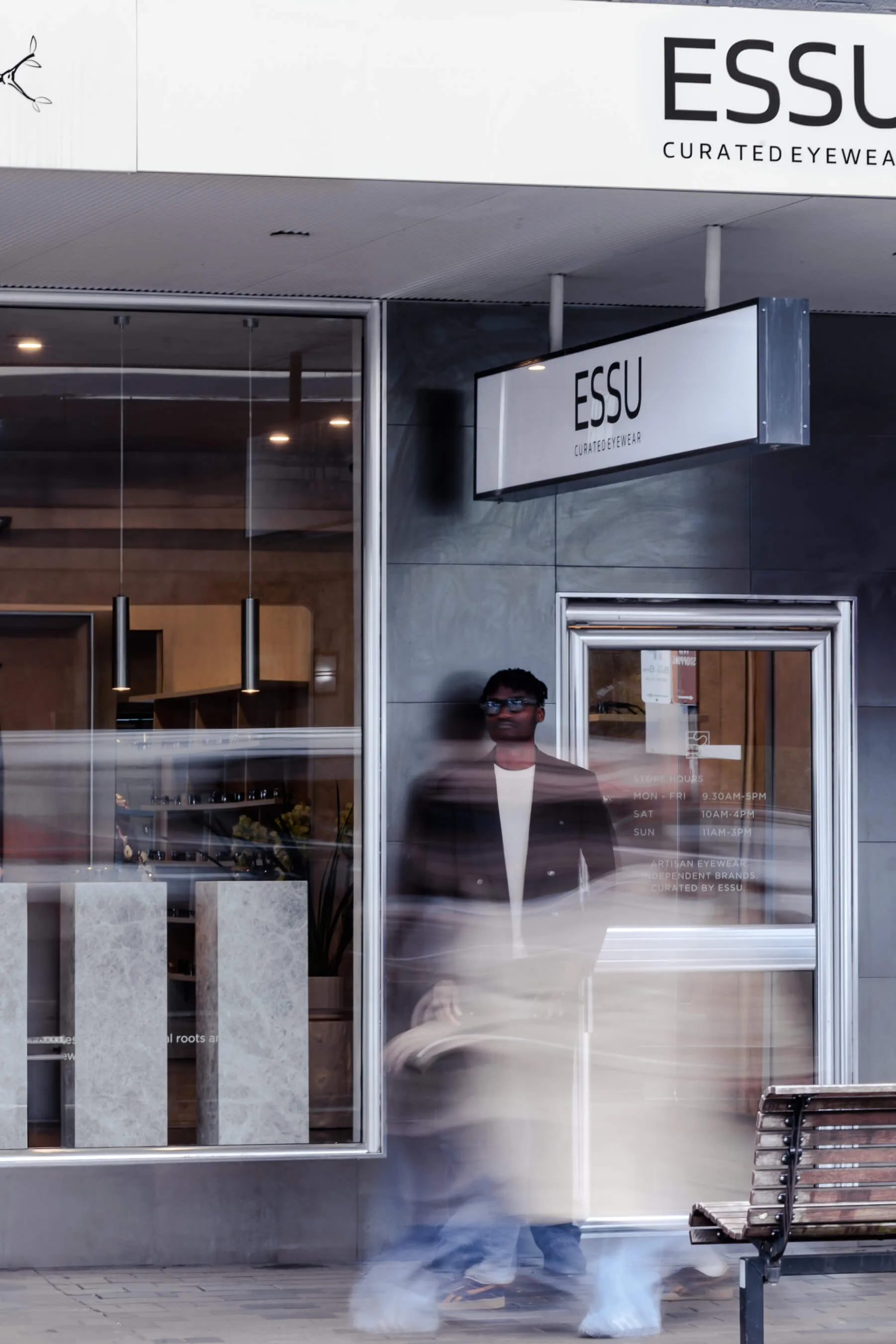 Long-exposure capture of the same storefront, with motion-blurred pedestrians walking past the model. ESSU’s branding, store hours, and the clean, modern design of the façade are clearly visible | EDG Studios
