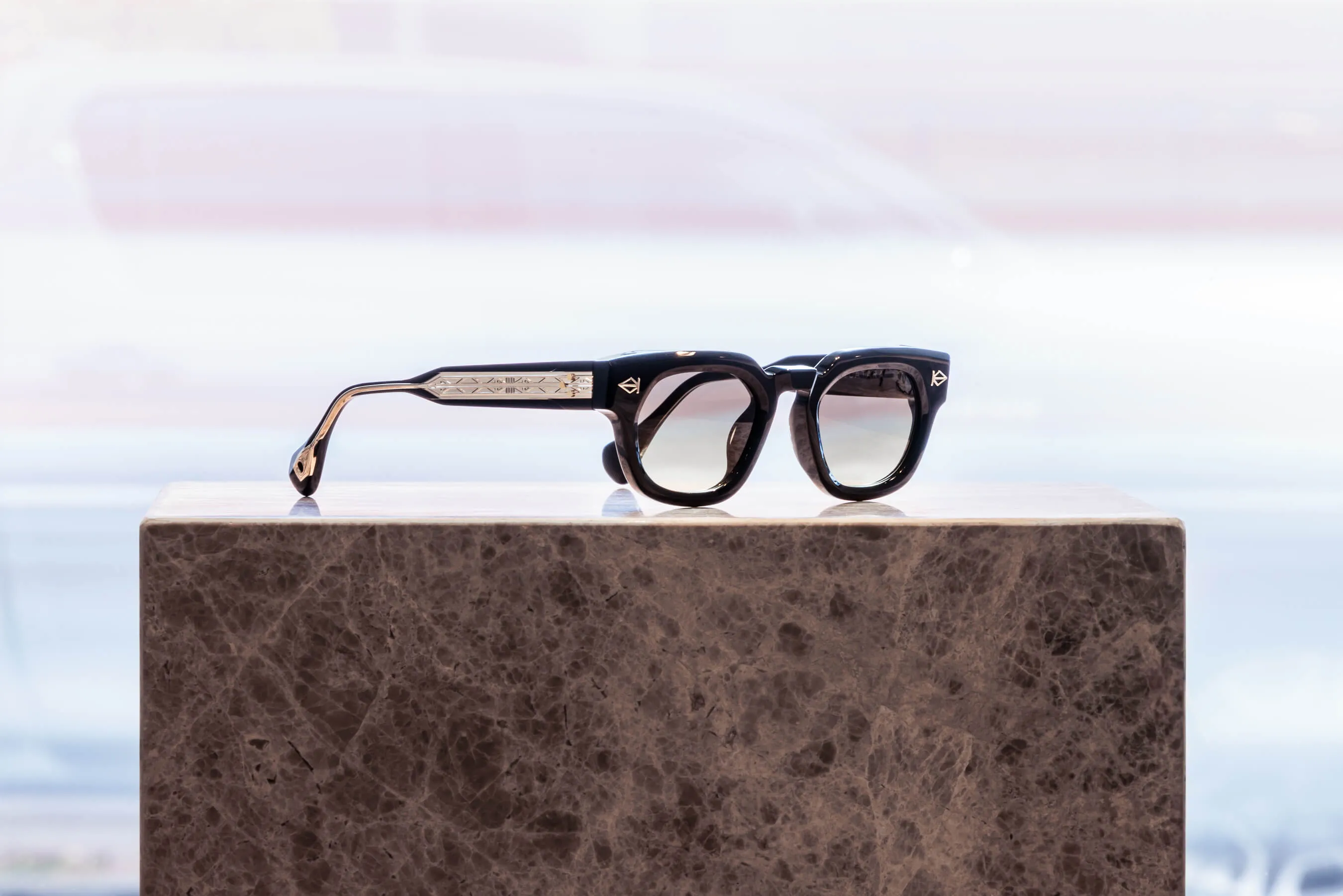 Bold, black acetate sunglasses with engraved metal detailing showcased at Essu in Mosman. The eyewear sits on a stone block, with natural daylight flooding through the storefront window behind | EDG Studios