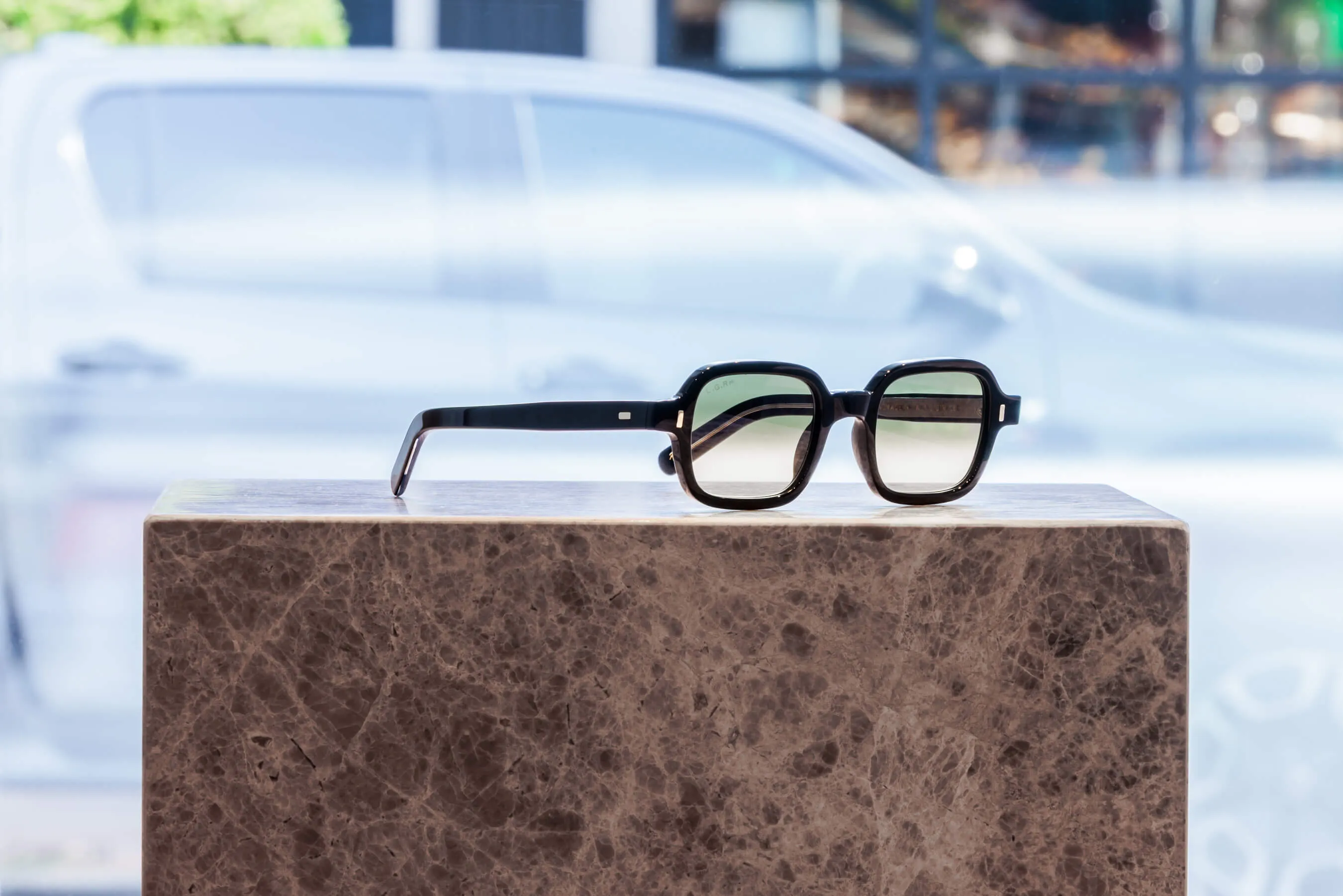 A modern, minimalist black sunglass frame with rounded square lenses, shown in soft daylight at Essu’s Mosman boutique. Positioned on polished stone with vehicle reflections outside the store window | EDG Studios