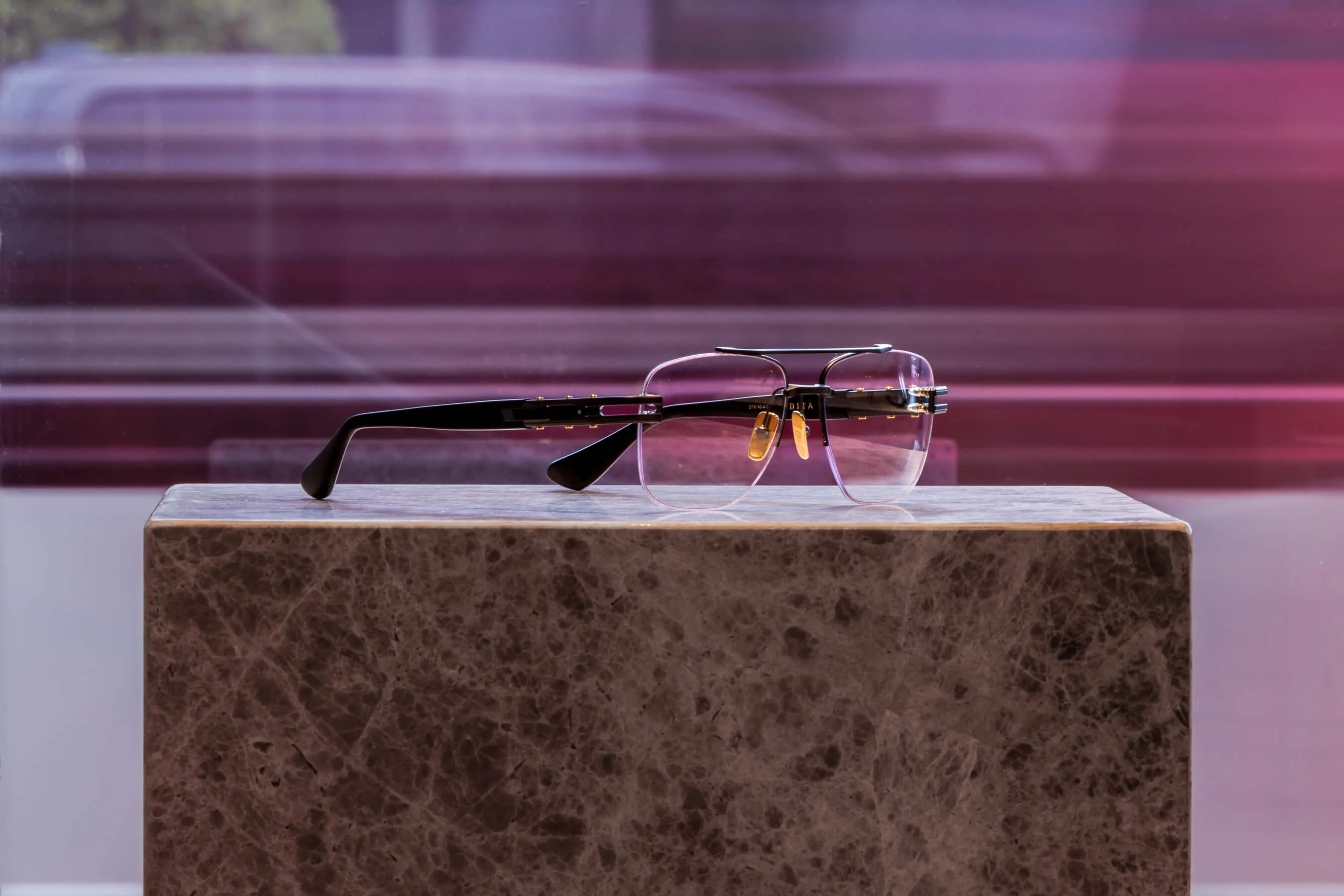 A pair of high-end Dita eyeglasses with gold accents and a rimless frame, set on a polished marble pedestal. Photographed at Essu Curated Eyewear in Mosman, Sydney, with a softly blurred city street in the backdrop | EDG Studios