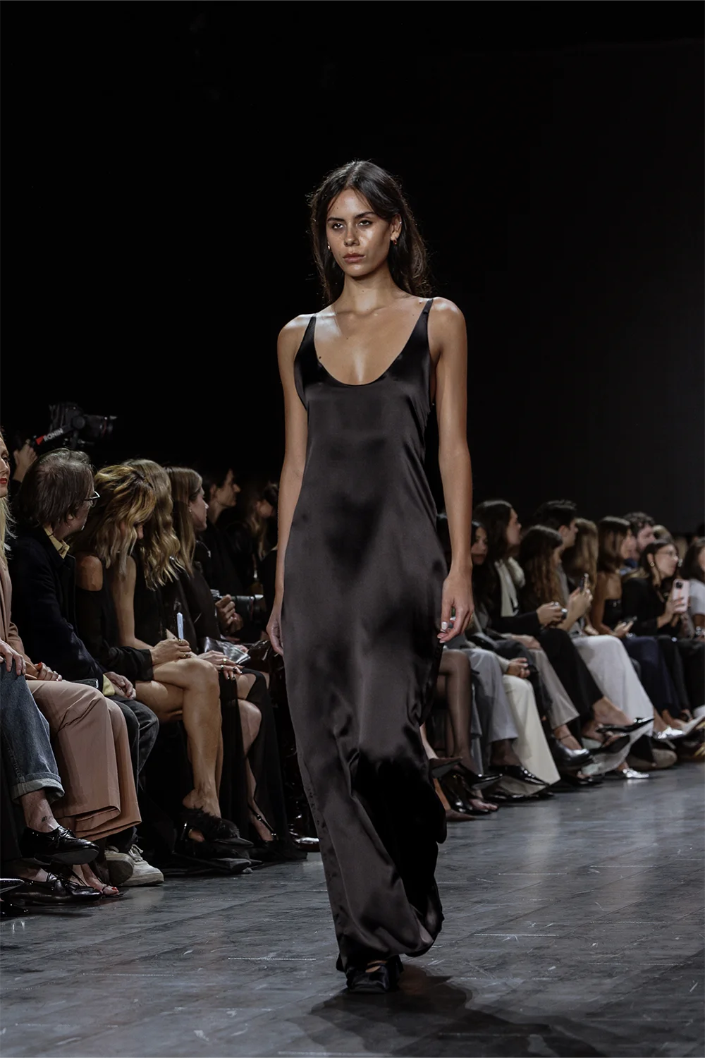 Full-length runway shot of a model in a floor-grazing, sleeveless black satin slip dress with a sleek silhouette and understated elegance.
