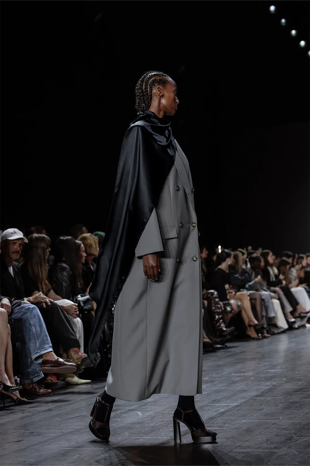 Runway side profile showing a model in a structured grey double-breasted overcoat with a black satin scarf draped across one shoulder.