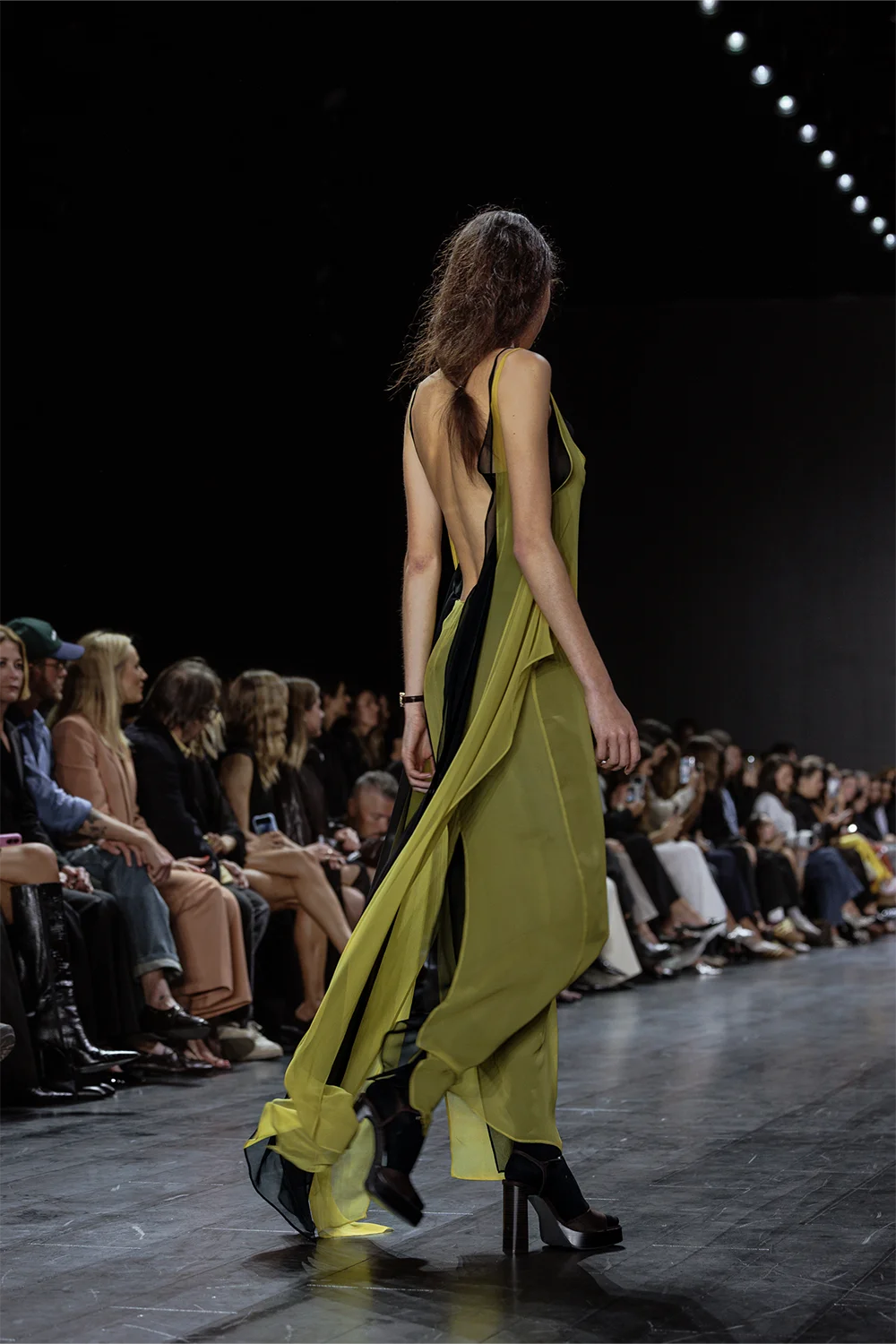 Model wearing a flowing, backless lime green chiffon overlay dress with a black inner slip, photographed from behind mid-walk on the runway.