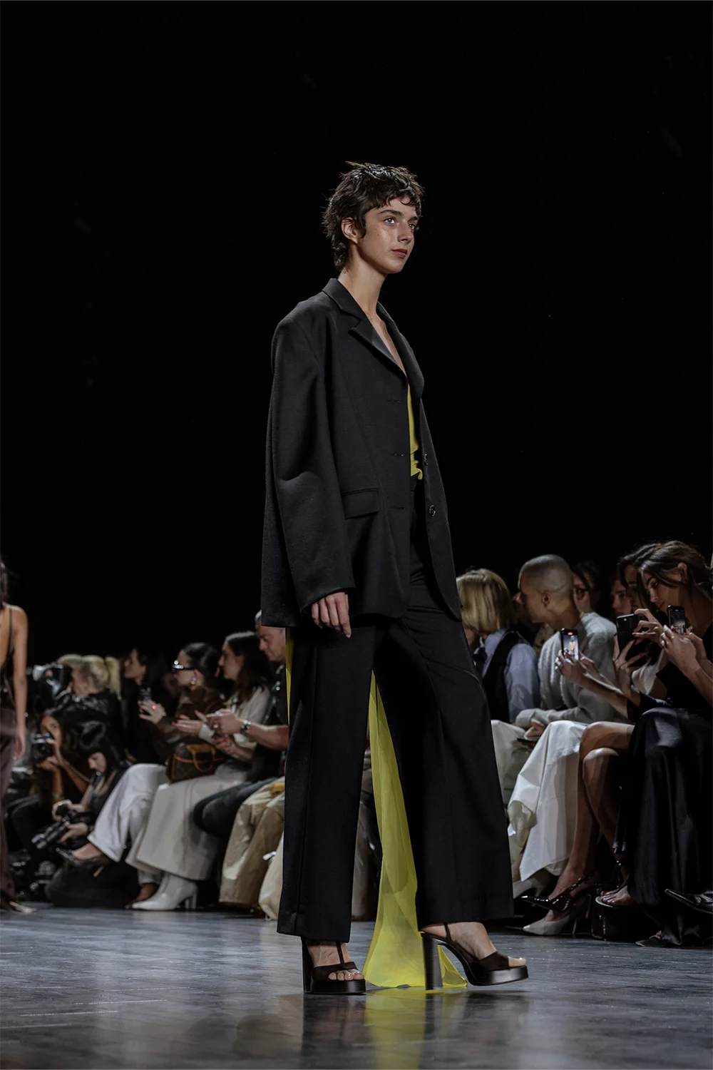 Model walking in a loose-fitting black pantsuit with wide-leg trousers, revealing a flowing lime chiffon inner panel beneath the blazer.