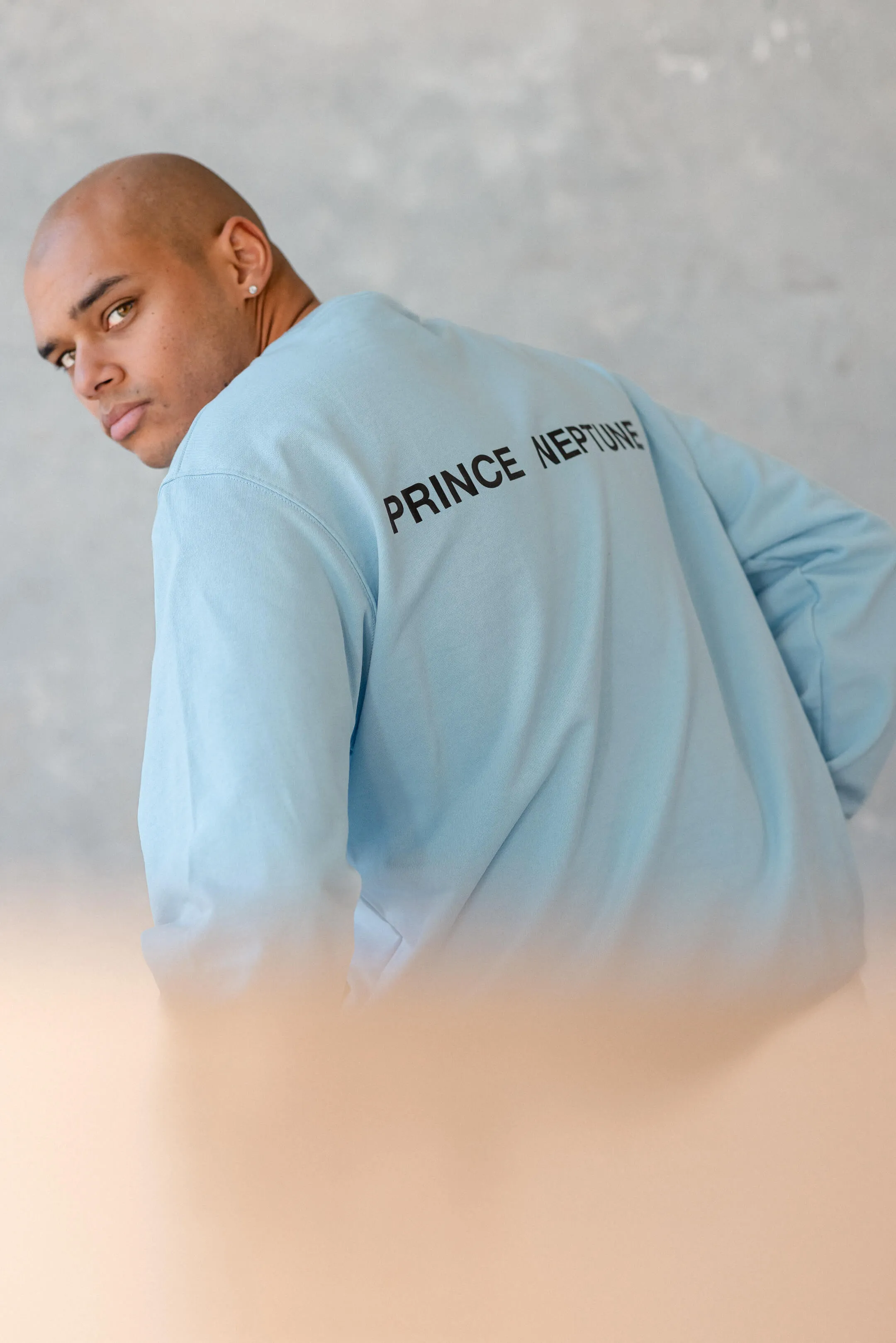 Model in sky blue Prince Neptune sweatshirt, looking over shoulder — A rear portrait of the model in a sky blue long sleeve shirt with “Prince Neptune” printed across the back, from the 2023 Collection One streetwear campaign | EDG Studios