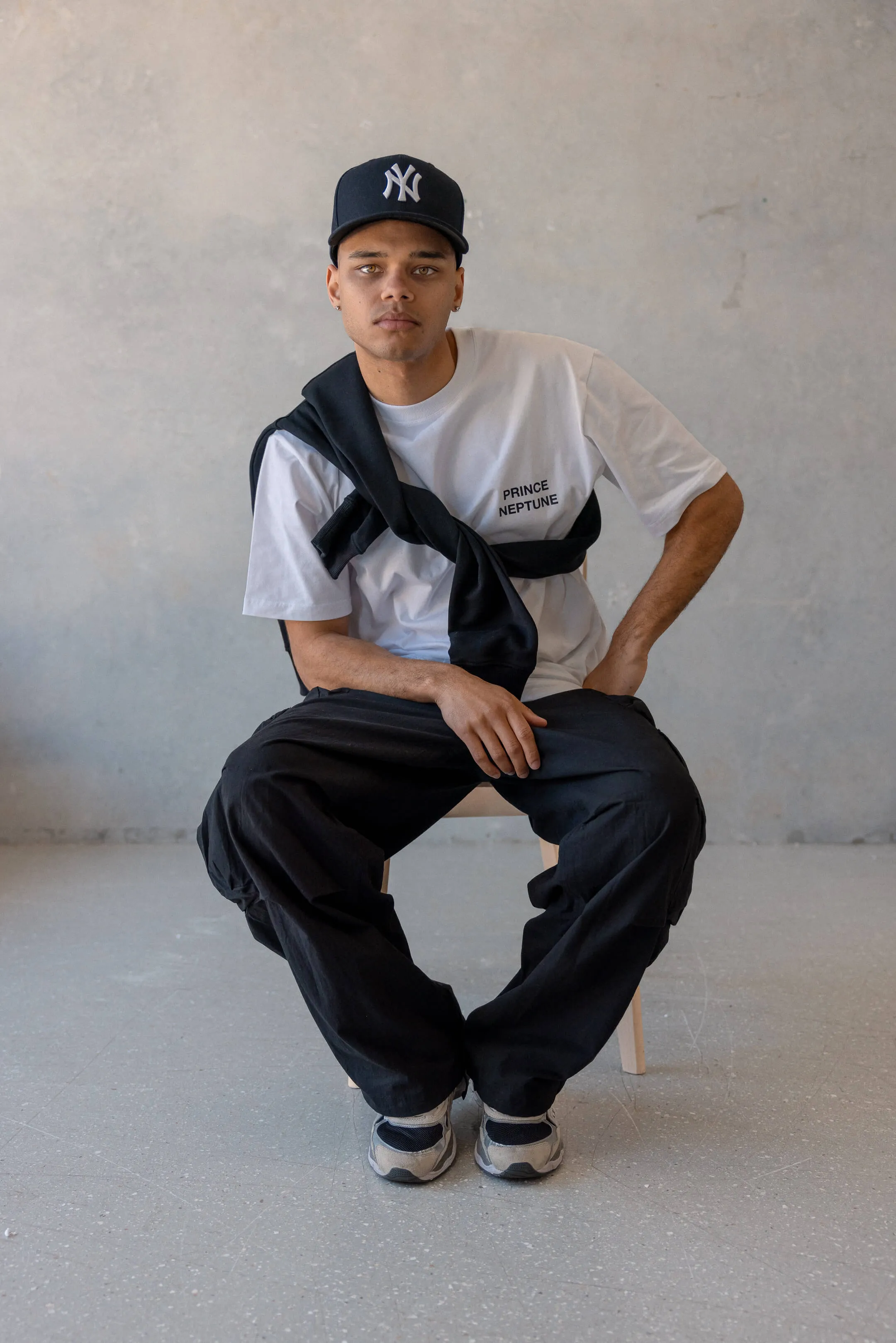 Model sitting in white Prince Neptune tee with jumper tied across chest — Seated pose of the model in a white Prince Neptune t-shirt with a black jumper tied across the shoulders, from Collection One, 2023 streetwear shoot | EDG Studios