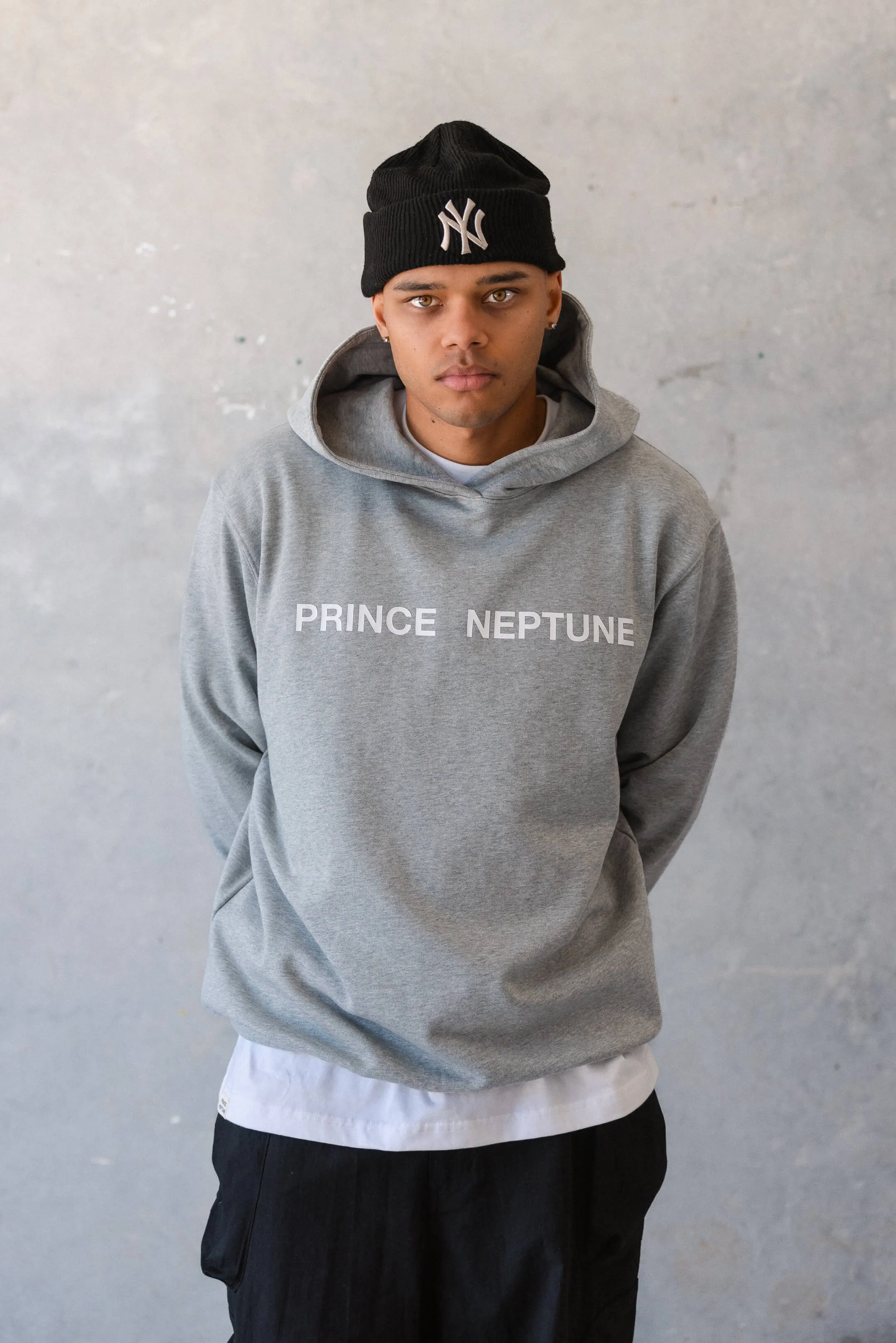Model in grey hoodie standing against wall — Full-length image of a model in a grey Prince Neptune hoodie layered over a white tee, styled for the 2023 Collection One streetwear campaign | EDG Studios