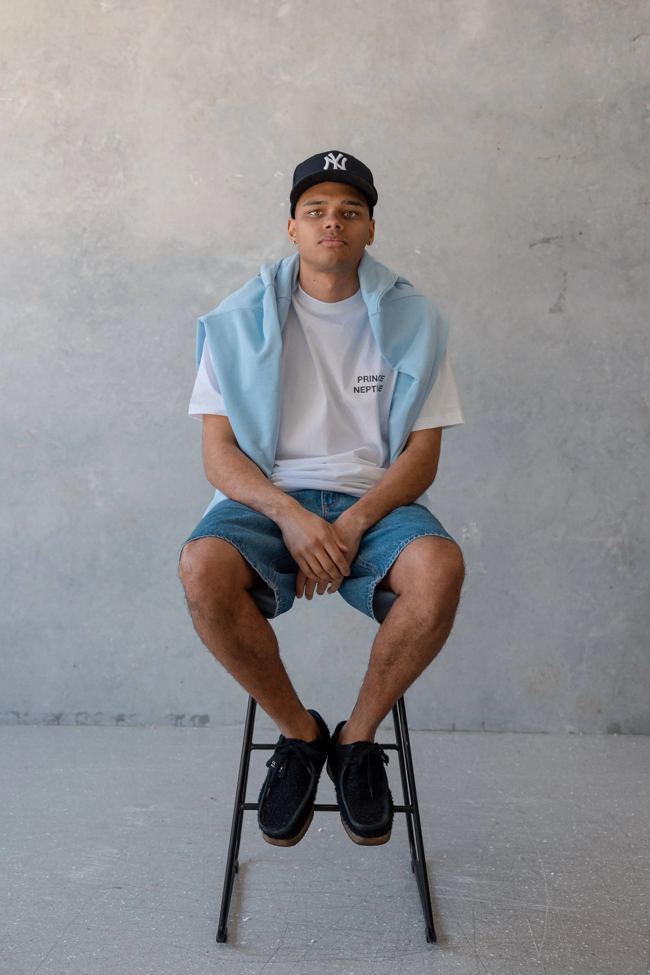 Model seated in light blue styled tee and denim shorts — Portrait of the model wearing a white Prince Neptune t-shirt and light denim shorts with a sky blue jumper, photographed for Collection One | EDG Studios