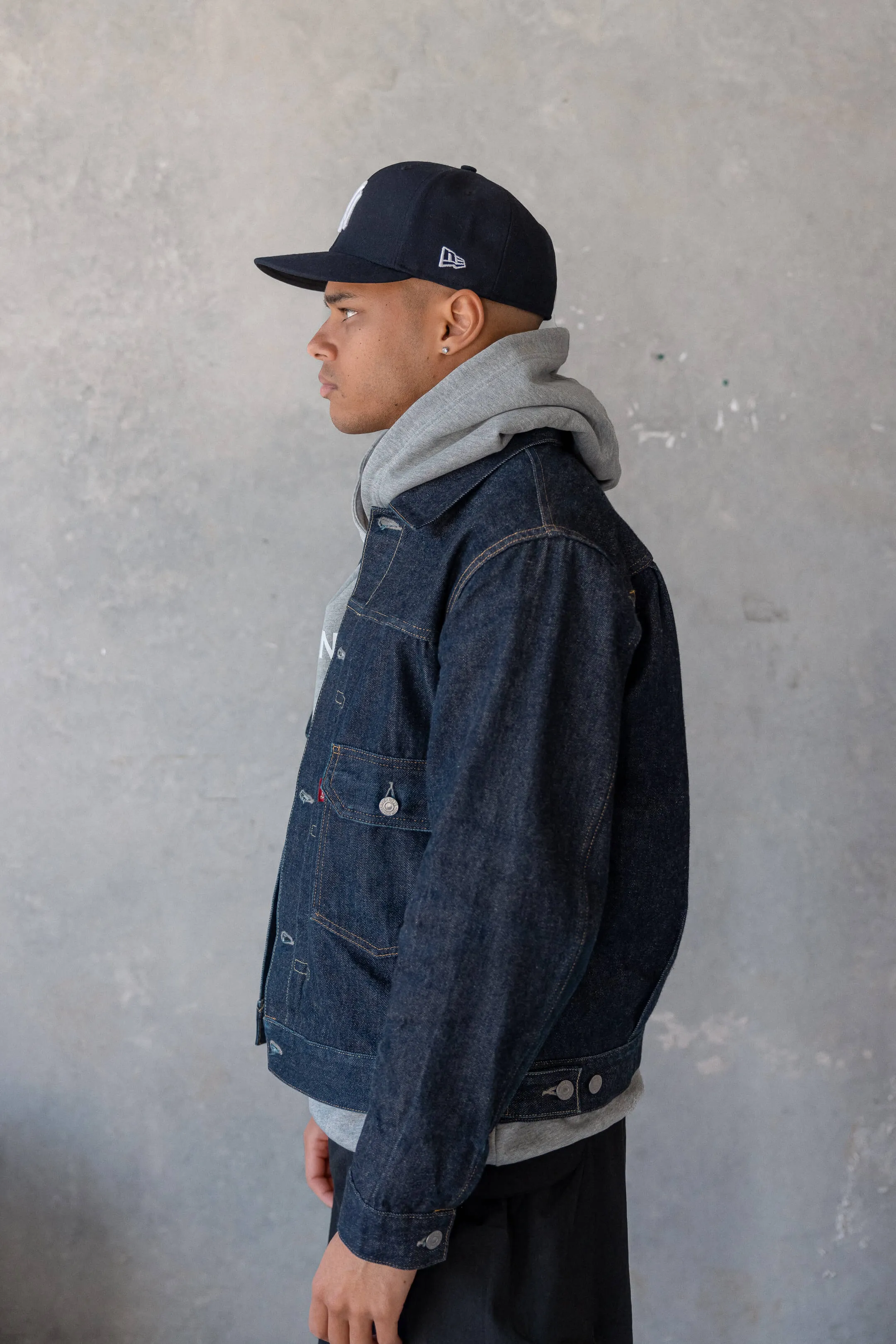 Model in denim jacket and grey hoodie — Side profile of a male model wearing a New York Yankees cap, layered denim jacket, and grey hoodie from the 2023 Prince Neptune Collection One streetwear lookbook | EDG Studios