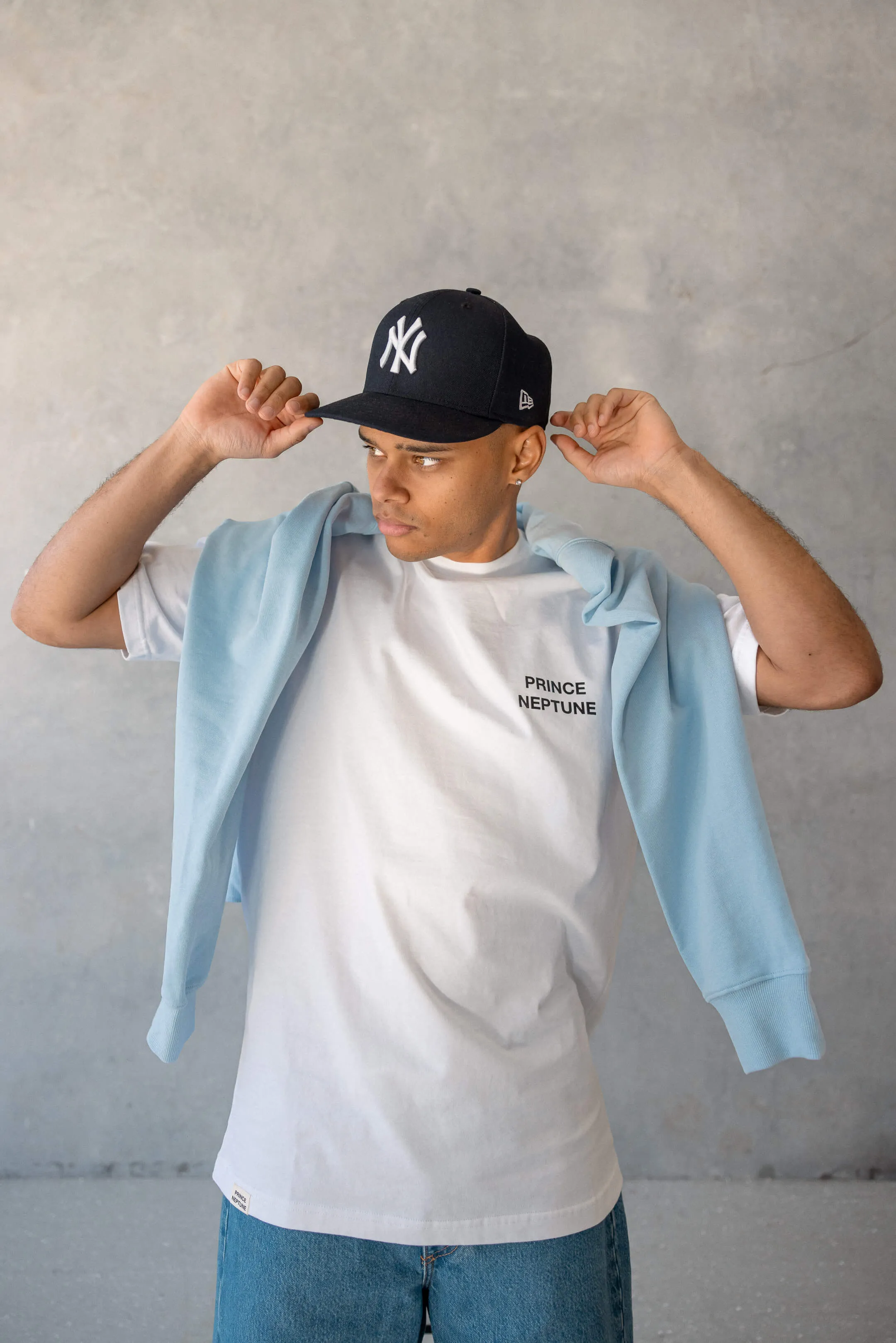 Model adjusting New York Yankees cap while wearing white Prince Neptune T-shirt and light blue sweater over shoulders, standing against a concrete wall | EDG Studios