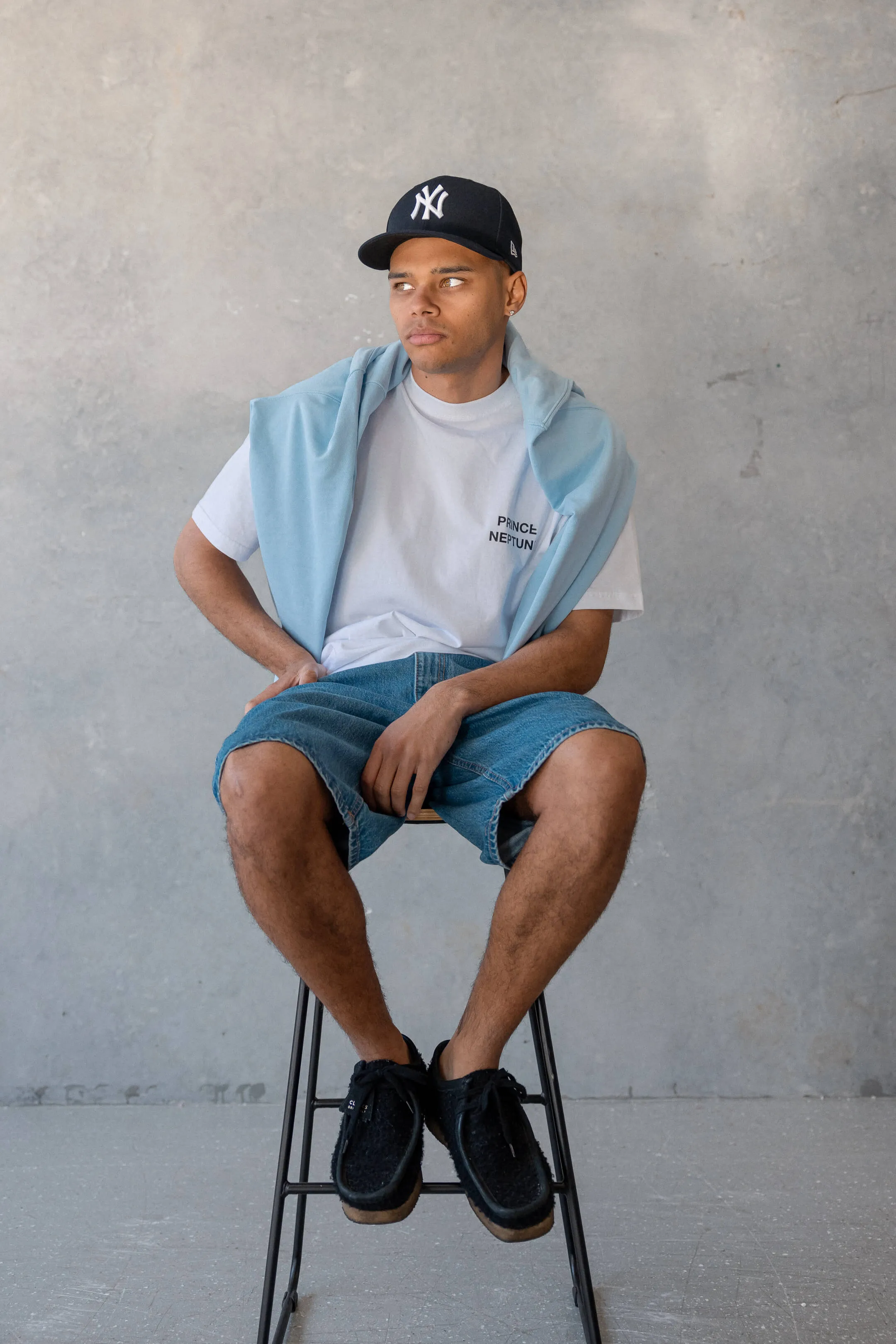 Model in casual pose with tee and blue jumper styled over shoulders — Editorial image of a model in a relaxed pose wearing a Prince Neptune tee and denim shorts, from the 2023 streetwear Collection One series | EDG Studios