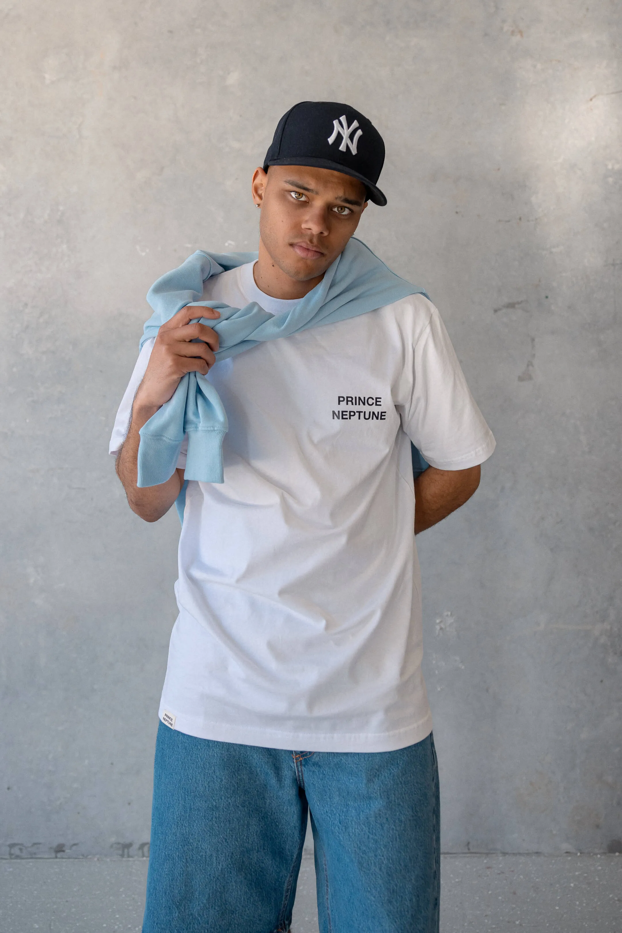 Model in Prince Neptune tee with sky blue jumper over shoulders — A portrait of a male model wearing a white “Prince Neptune” t-shirt and denim shorts, styled with a sky blue jumper over the shoulders, from the 2023 streetwear shoot for Prince Neptune Collection One | EDG Studios