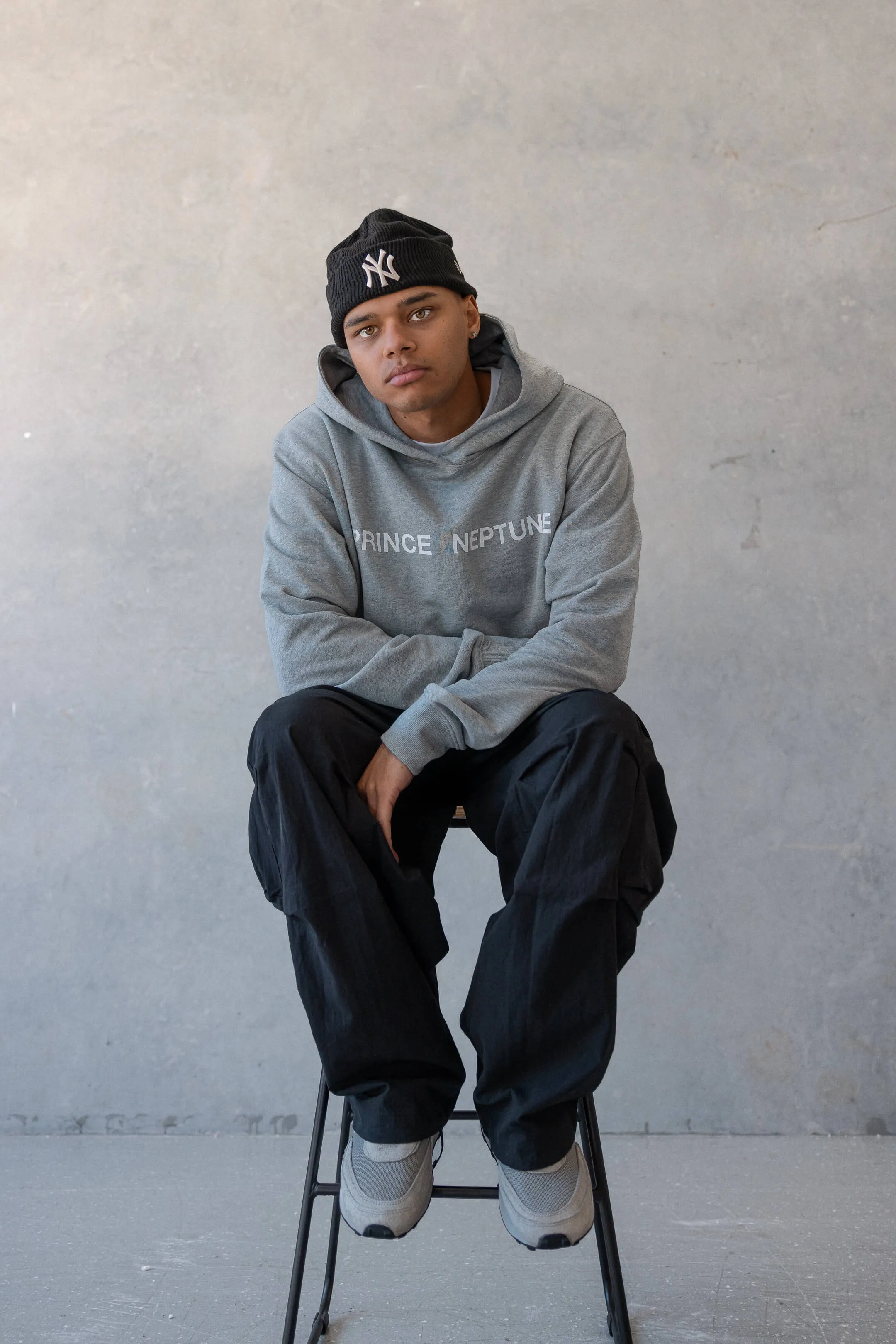 Model seated on stool in grey hoodie and black beanie — A relaxed editorial portrait featuring the model in a Prince Neptune hoodie and cargo pants, captured for Collection One, 2023 | EDG Studios