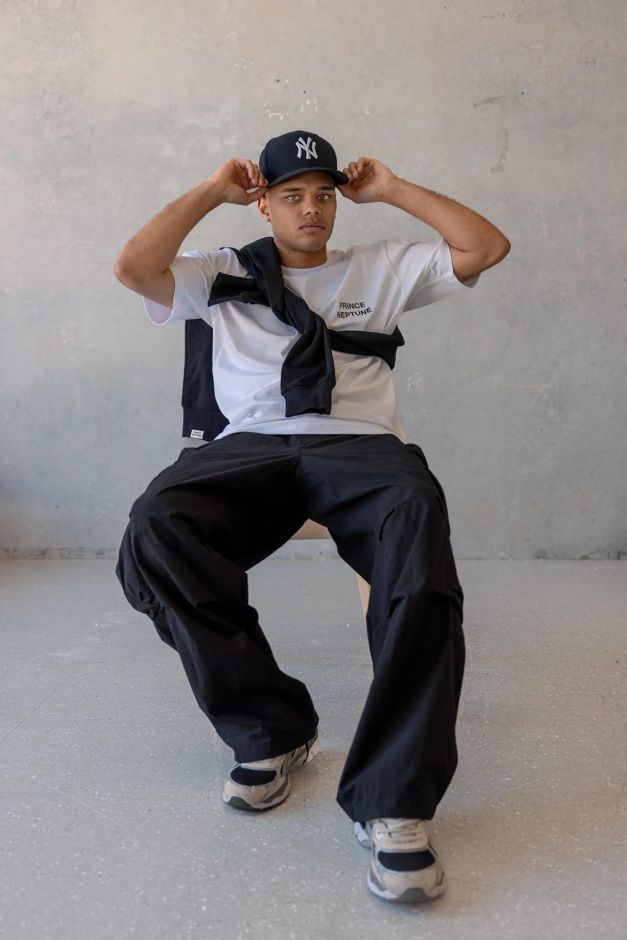 Model seated and tipping Yankees cap, styled in Prince Neptune tee, black cargo pants, and black sweater draped over shoulders | EDG Studios