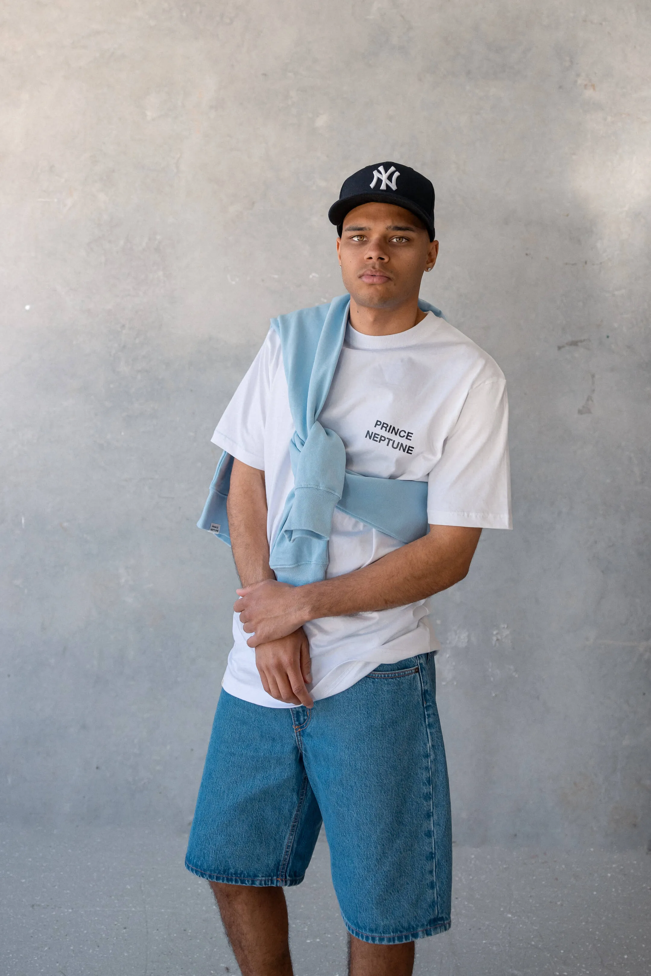 Model standing front-facing with arms crossed, styled in Prince Neptune white tee and tied blue sweater, denim shorts, and Yankees cap | EDG Studios