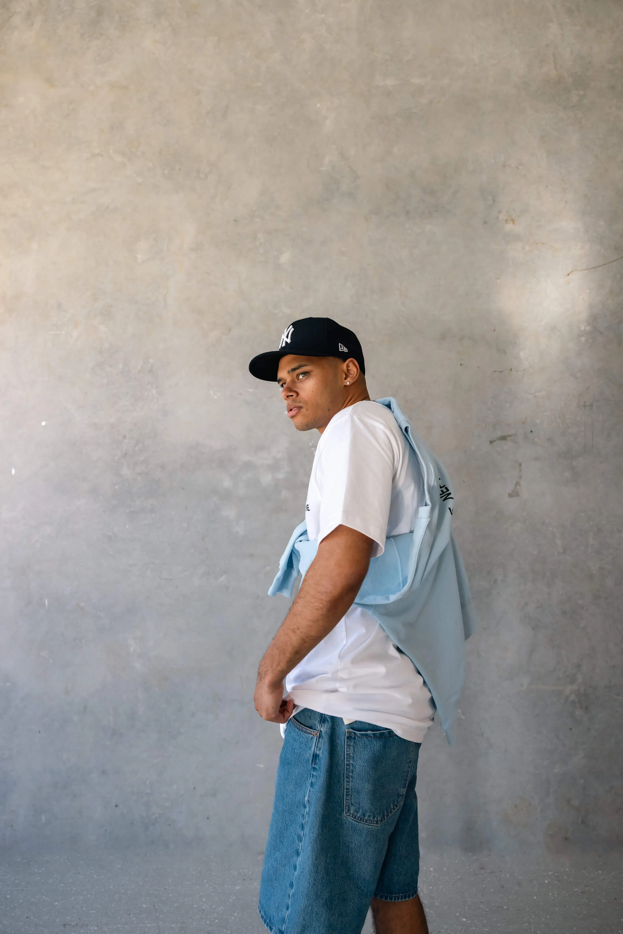 Side profile of model in Prince Neptune tee and blue denim shorts, looking over shoulder with light blue sweater draped over back | EDG Studios