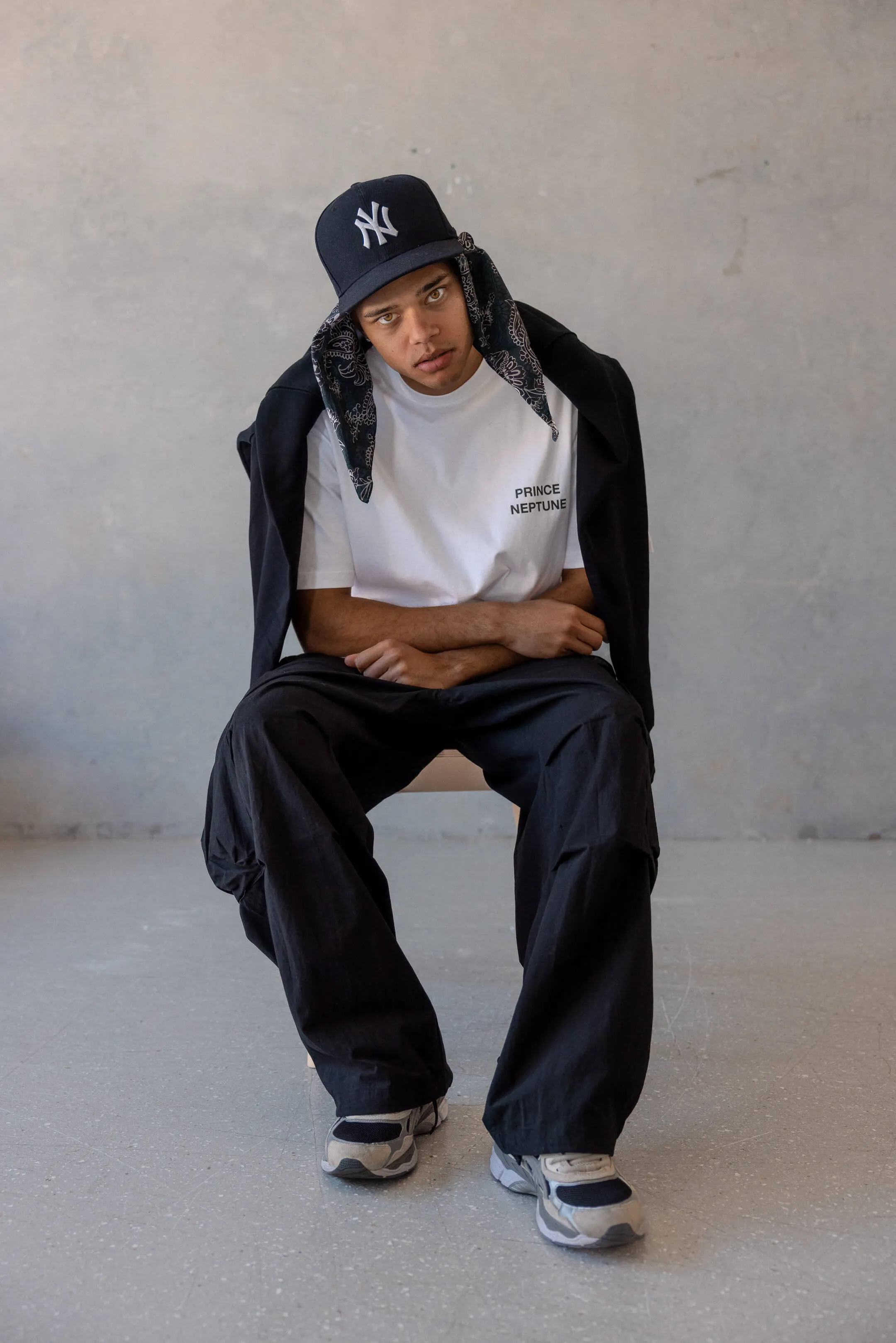 Seated model wearing Prince Neptune tee, black trousers, sneakers, and black jacket draped over shoulders with paisley bandana beneath cap | EDG Studios