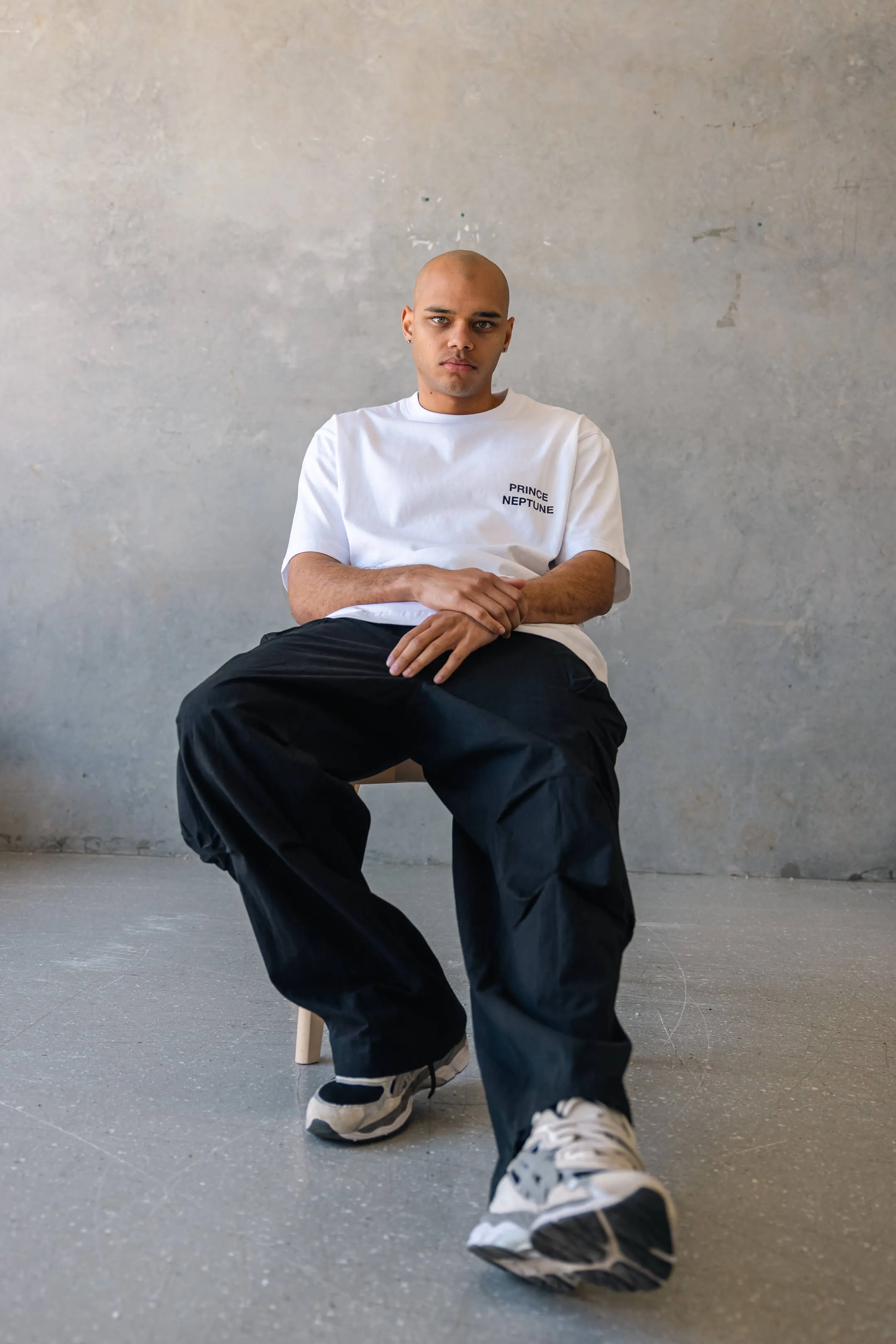 Model seated on stool in relaxed pose, wearing white Prince Neptune T-shirt, black cargo pants, and sneakers | EDG Studios